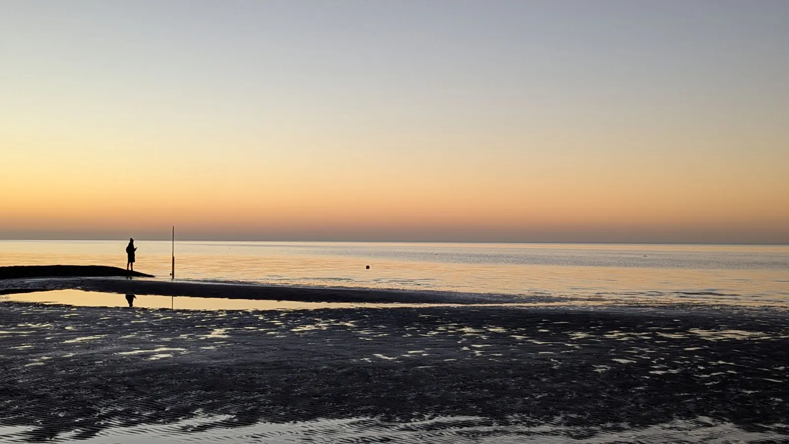 Ein Bild, das ich letzte Woche in Cuxhaven am Sahlenburger Strand geschossen habe. Man sieht den Strand bei Abenddämmerung, der Himmel leuchtet in rötlichen Farben, im Vordergrund noch ein wenig Watt, weiter hinten kehrt bereits die Flut zurück. Ganz links, ganz klein die Silhouette einer Person, direkt am Wasser, die in die Ferne schaut.