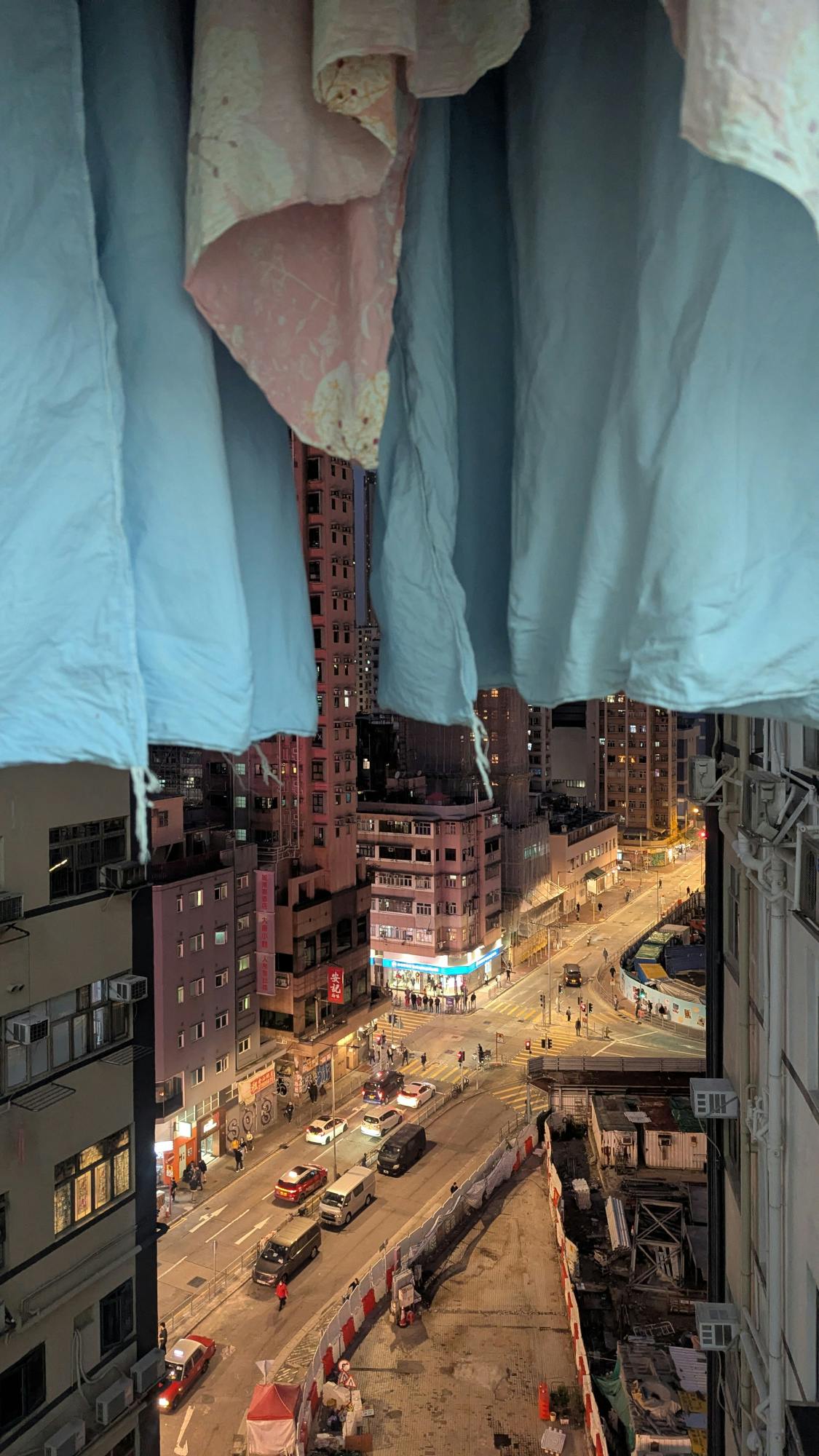 Unter hängender lichtblauer Bettwäsche öffnet sich der Blick auf eine nächtliche, geöb beleuchtete Straßenlandschaft tief unten. Wenige Autos, eine Baustelle, hohe Häuser, kaum Menschen.