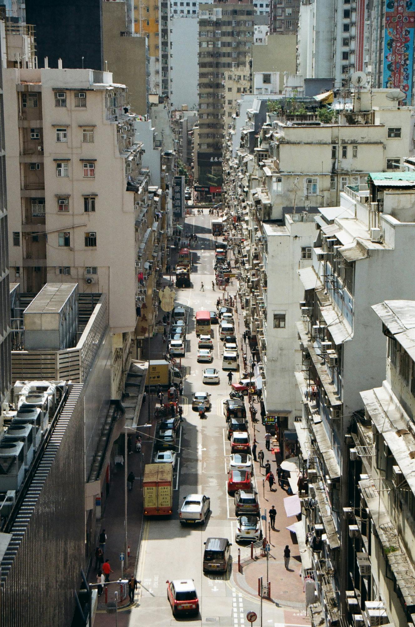 Blick in eine sonnenbeschienene Straßenschlucht zwischen terrassierten hohen Häusern, abgeschlossen von einer Wand noch höhere Häuser. Autos und Menschen die scharfe Schatten werfen.