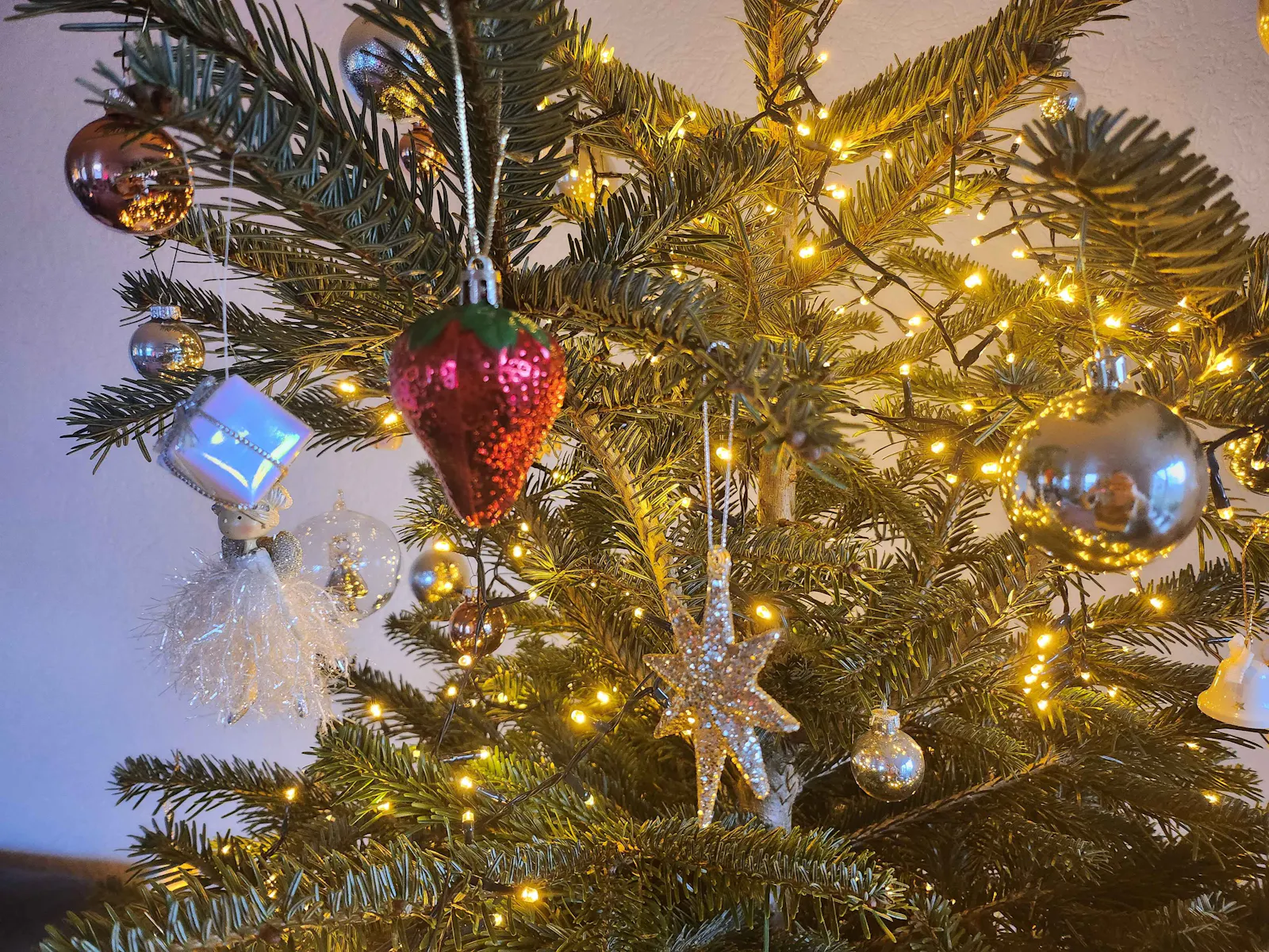 Nahaufnahme eines geschmückten Weihnachtsbaums mit warmweißen Lichterketten, goldenen und orangenen Christbaumkugeln sowie funkelnden Anhängern in Stern- und Engel-Form. Eine Erdbeere ist auch zu sehen.