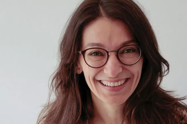 Porträt einer lachenden Person mit langen braunen Haaren und runder Brille vor einem hellen Hintergrund.