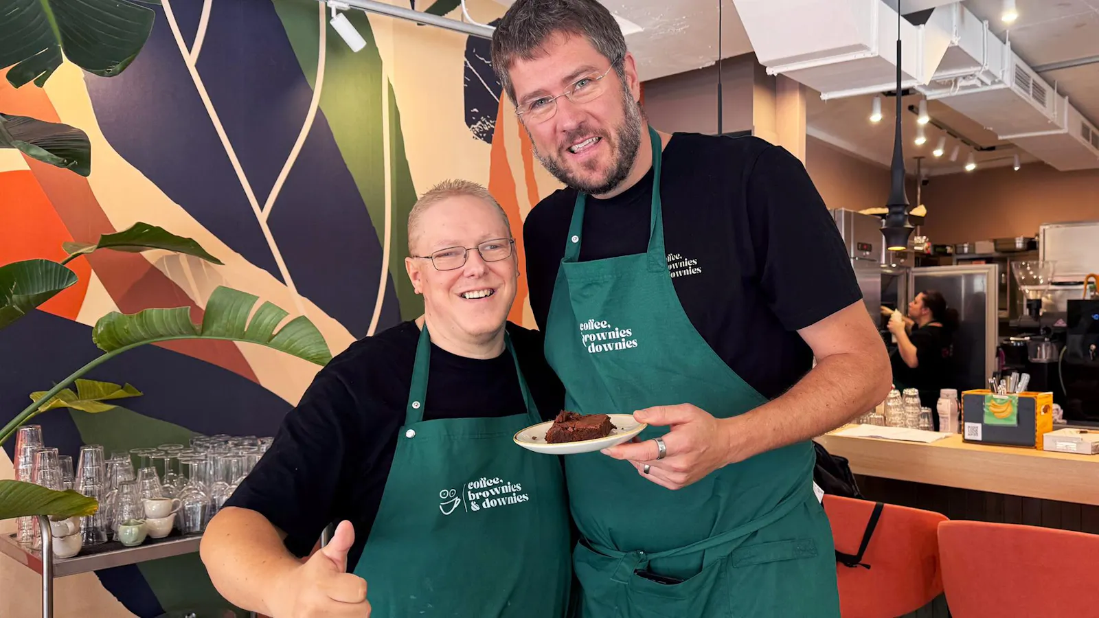 Zwei Männer mit grünen Gastronomieschürzen stehen dicht nebeneinander in einem Café und lachen in die Kamera. Einer hält einen Teller mit einem Brownie. Der andere streckt den Daumen nach oben.
