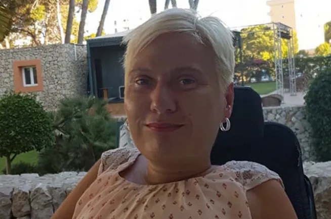 Porträt einer Person mit kurzen, hellblonden Haaren und Ohrringen, die draußen vor Steinmauern und Bäumen sitzt und in die Kamera lächelt.