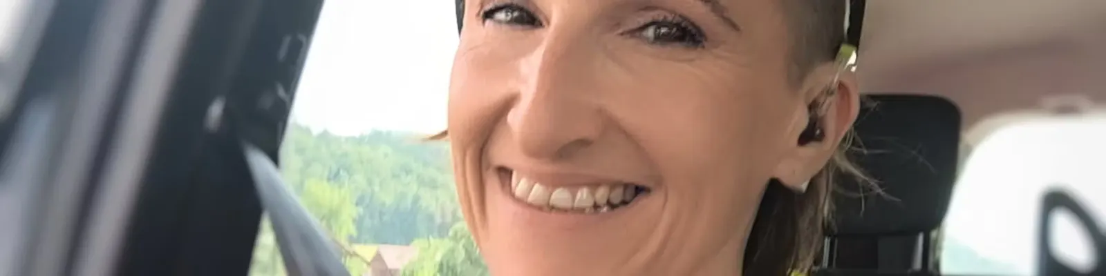 Nahaufnahme einer lächelnden Person mit kurzem Haar und Ohrhörern, die im Auto sitzt. Durch das Fenster ist eine grüne Landschaft zu sehen.