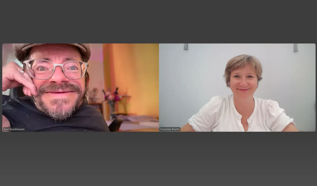 Screenshot eines Video-Calls mit zwei Personen. Links sitzt eine Person mit Brille, Bart und Mütze in einem warm beleuchteten Raum, rechts eine Person mit kurzem Haar im weißen Oberteil vor hellem Hintergrund.