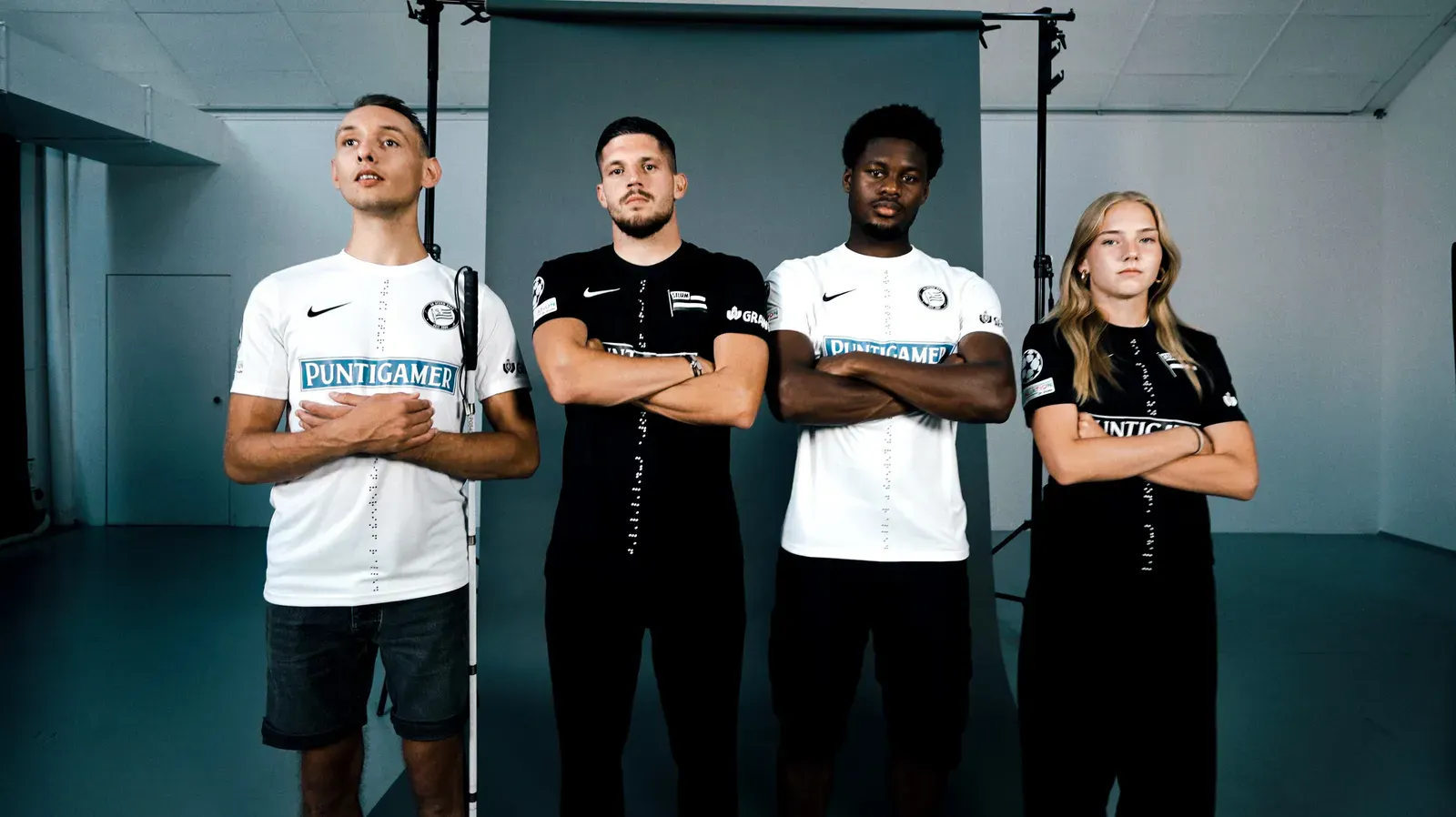Vier Fußballspielerinnen und -spieler stehen nebeneinander vor grauem Hintergrund und posieren mit verschränkten Armen. Zwei tragen weiße Trikots mit der Aufschrift „Puntigamer“, zwei schwarze Trikots desselben Designs.