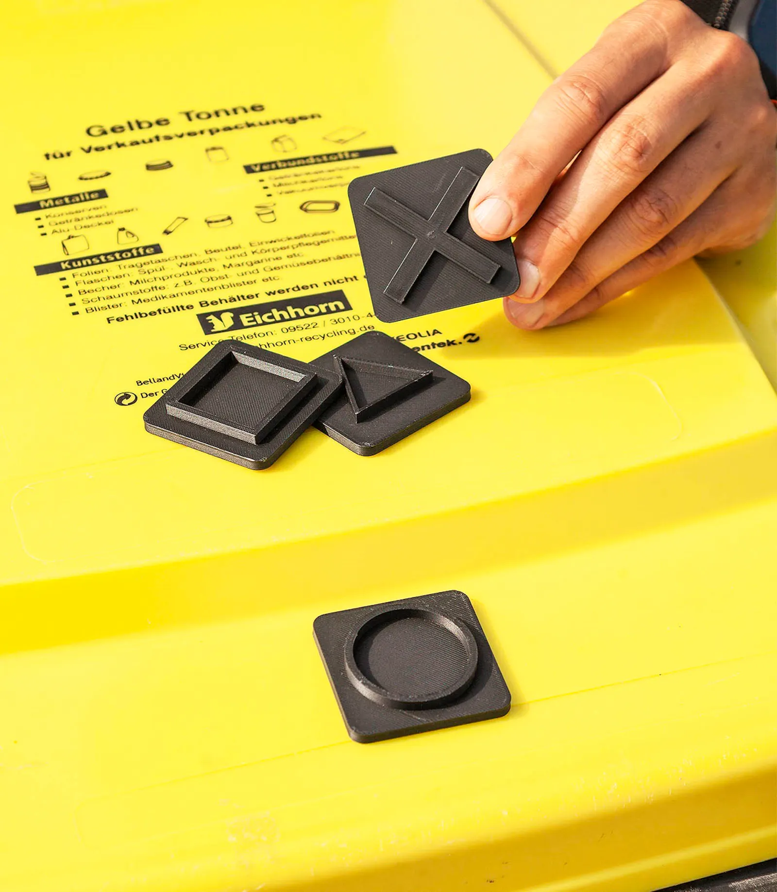 Eine Hand legt ein schwarzes Kunststoffteil auf den Deckel einer gelben Tonne für Verkaufsverpackungen. Auf dem Deckel liegen mehrere kleine schwarze Kunststoffelemente in unterschiedlichen Formen.