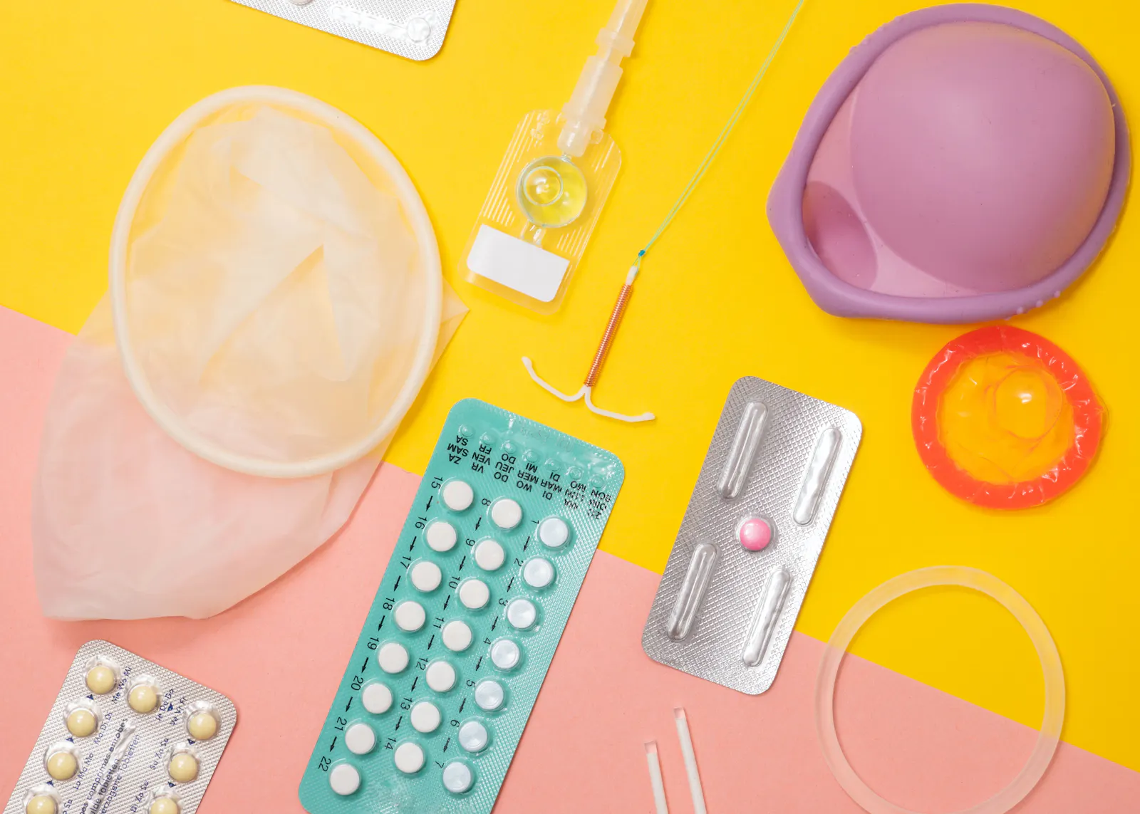 Verschiedene Verhütungsmittel, darunter die Pille in einem Blister, Diaphragma oder Kondom auf einem pink-gelbem Untergrund.