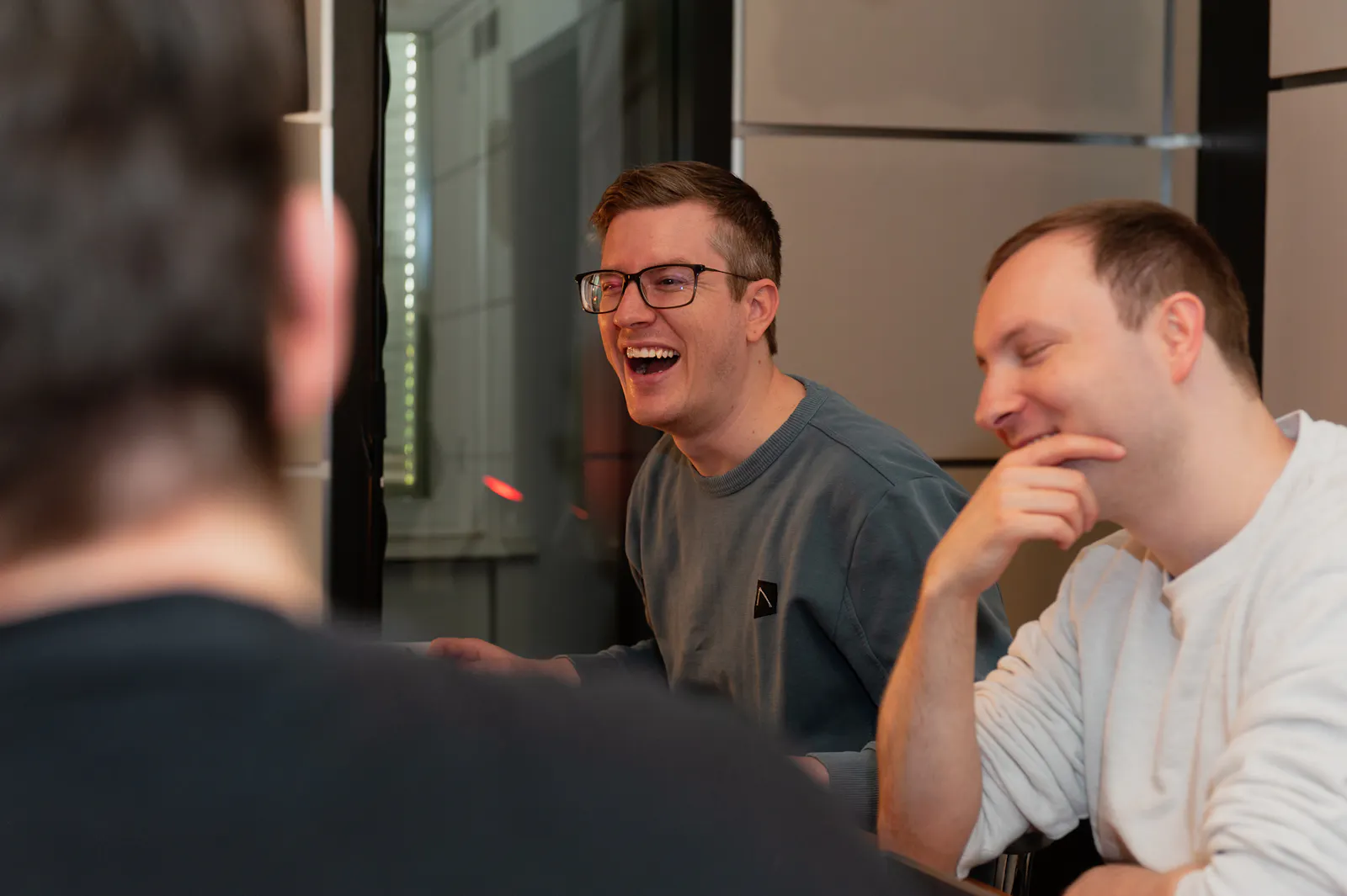 Tim sitzt am Spieltisch. Sein Mund ist weit lachend geöffnet. Neben ihm sitzt Kelly, ebenfalls schmunzelnd.