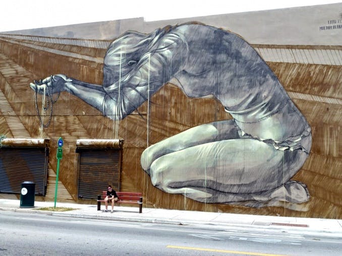 StreetArt der Künstlerin Faith47, fotografiert in Los Angeles. Das Graffiti zeigt eine junge Frau von der Seite in Grautönen. Sie trägt einen Slipp und ein knappes langärmeliges Oberteil. Sie hockt auf ihren Unterschenkeln, den Kopf, mit langen geraden Haaren, zwischen den Armen verborgen. Die Arme und Hände hat sie ausgestreckt und bietet einem nicht sichtbaren Gegenüber einige Ketten / Bänder dar.