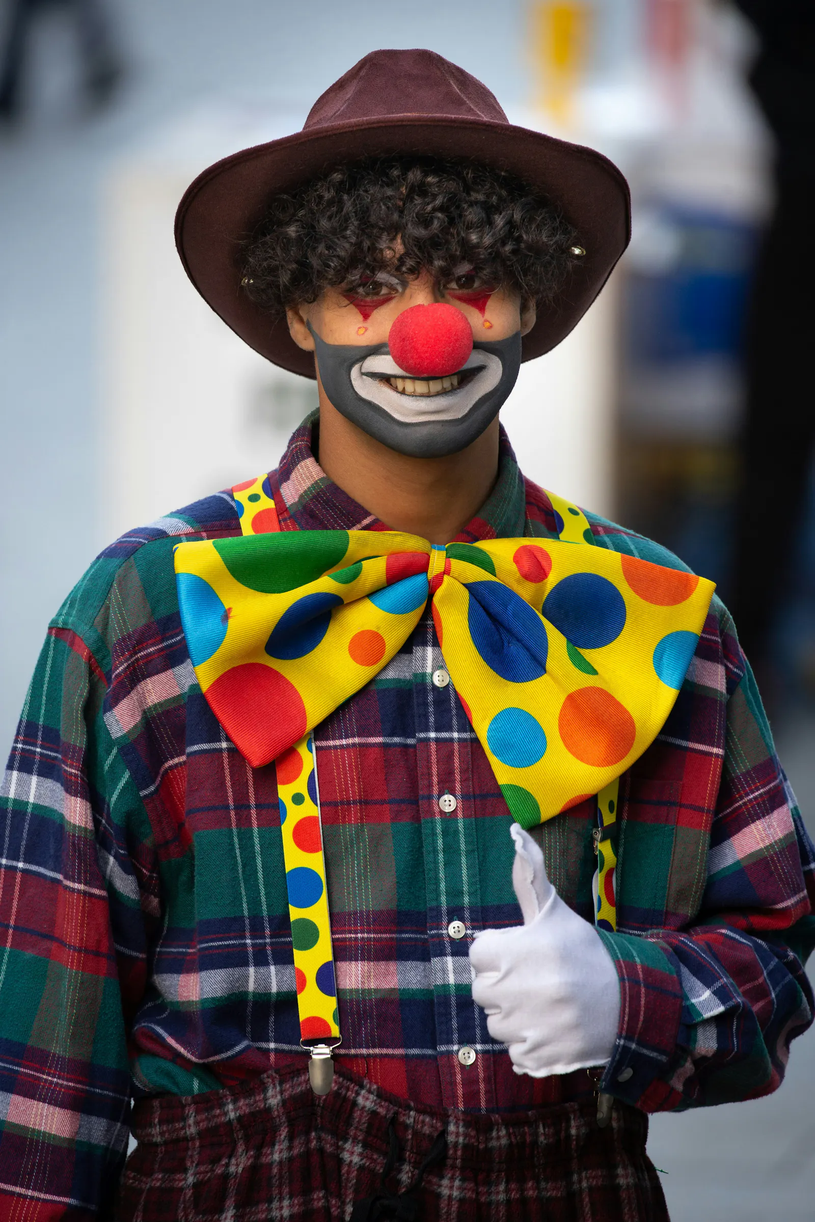Foto eines Menschen, der als Clown verkleidet ist. Er trägt einen dunklen Hut, hat eine rote Clownsnase und eine riesige bunte Fliege. Seine linke Hand zeigt die Daumen hoch-Geste