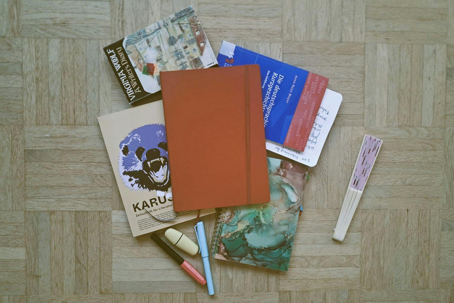 Verschiedene Notizbücher, die Zeitschrift Karussell, das Buch A Writer's Diary von Virginia Woolf, Eine Einführung in die deutschsprachige Kurzgeschichte von Anne-Rose Meyer, Stifte und ein Fächer liegen auf dem Boden.