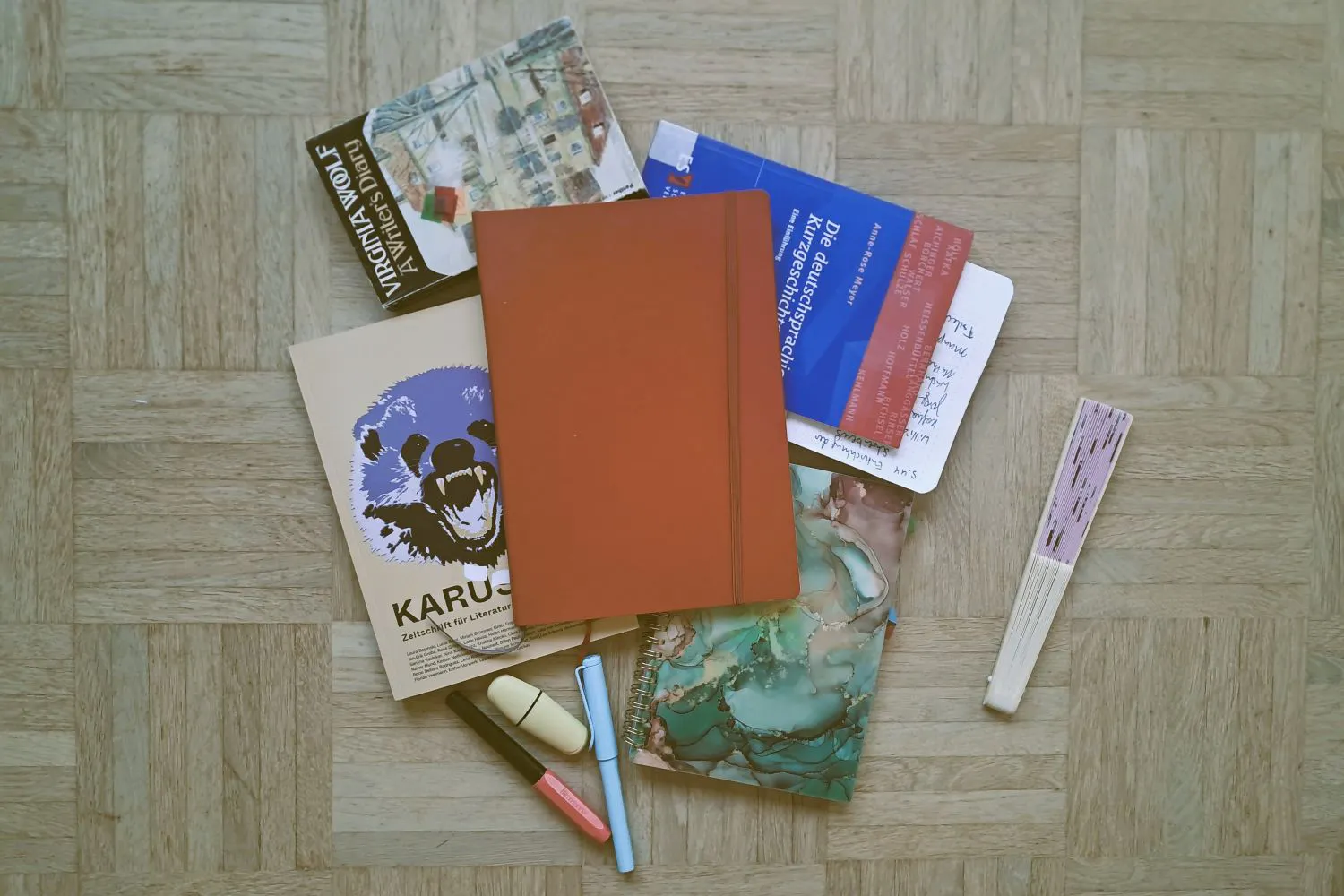 Verschiedene Notizbücher, die Zeitschrift Karussell, das Buch A Writer's Diary von Virginia Woolf, Eine Einführung in die deutschsprachige Kurzgeschichte von Anne-Rose Meyer, Stifte und ein Fächer liegen auf dem Boden.
