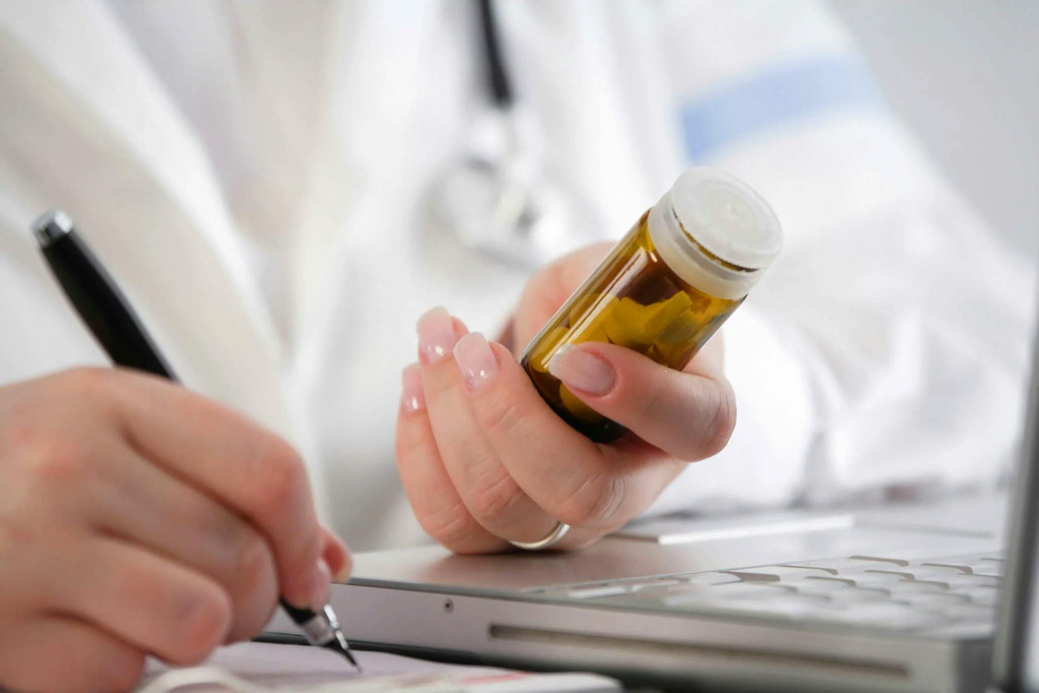 Die Hände einer Ärztin sind zu sehen, sie sitzt an einem Tisch, auf dem ein Laptop steht. Mit der einen Hand schreibt sie auf ein Papier neben dem Laptop, mit der anderen Hand hält sie ein gelbes Glasröhrchen, in dem Tabletten sind