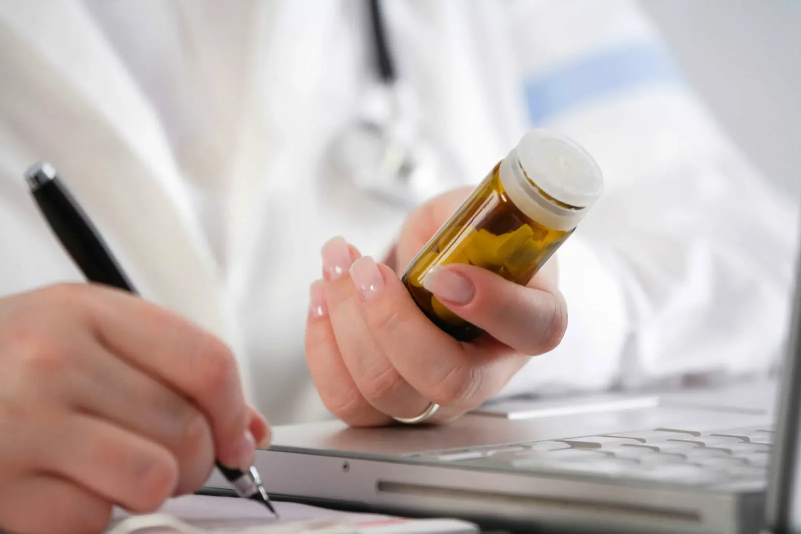 Die Hände einer Ärztin sind zu sehen, sie sitzt an einem Tisch, auf dem ein Laptop steht. Mit der einen Hand schreibt sie auf ein Papier neben dem Laptop, mit der anderen Hand hält sie ein gelbes Glasröhrchen, in dem Tabletten sind