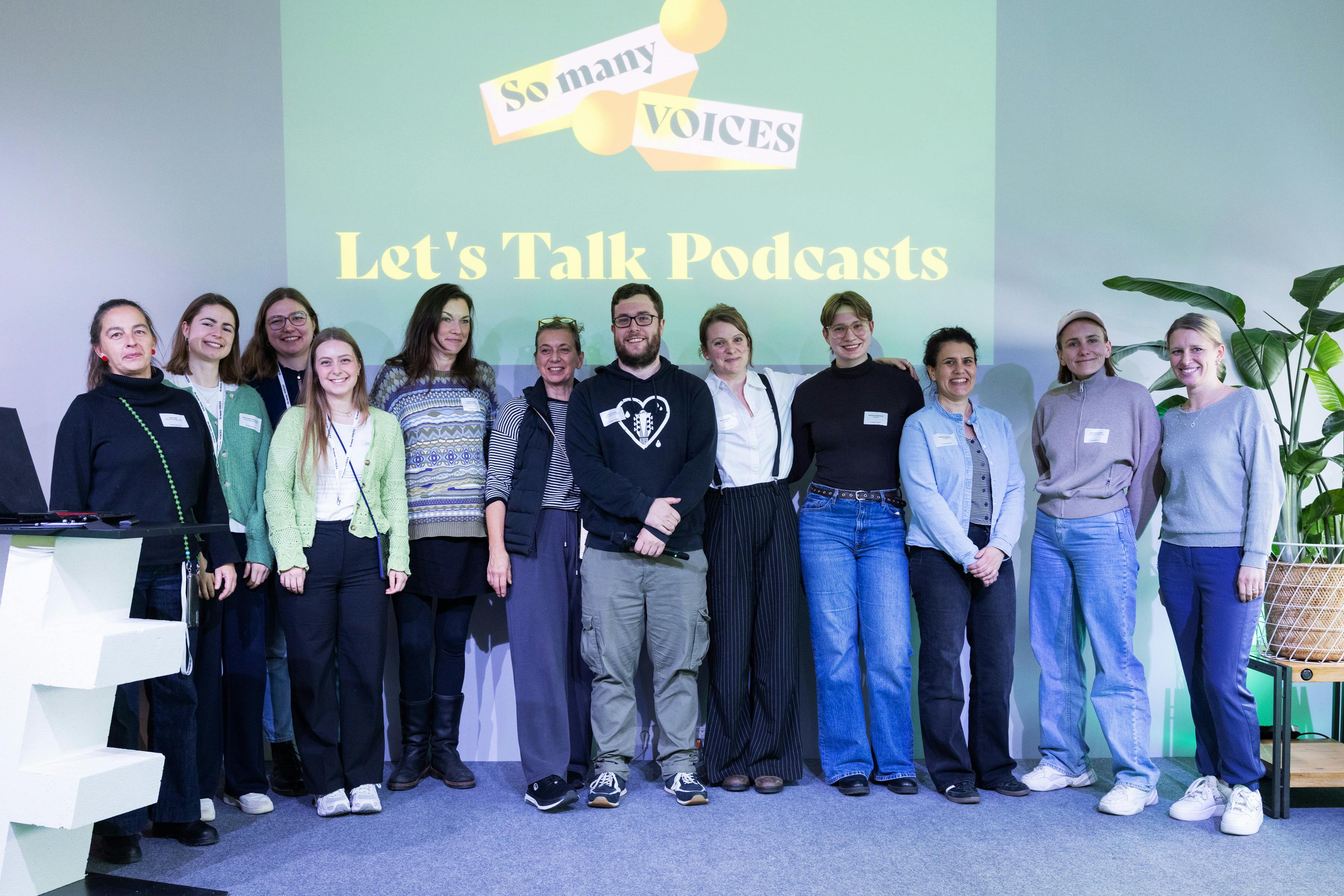 Eine Gruppe von Menschen steht auf der Bühne der So many Voices. Im Hintergrund ist das Logo der Konferenz zu sehen und die Aufschrift „Lets talk Podcast“.
