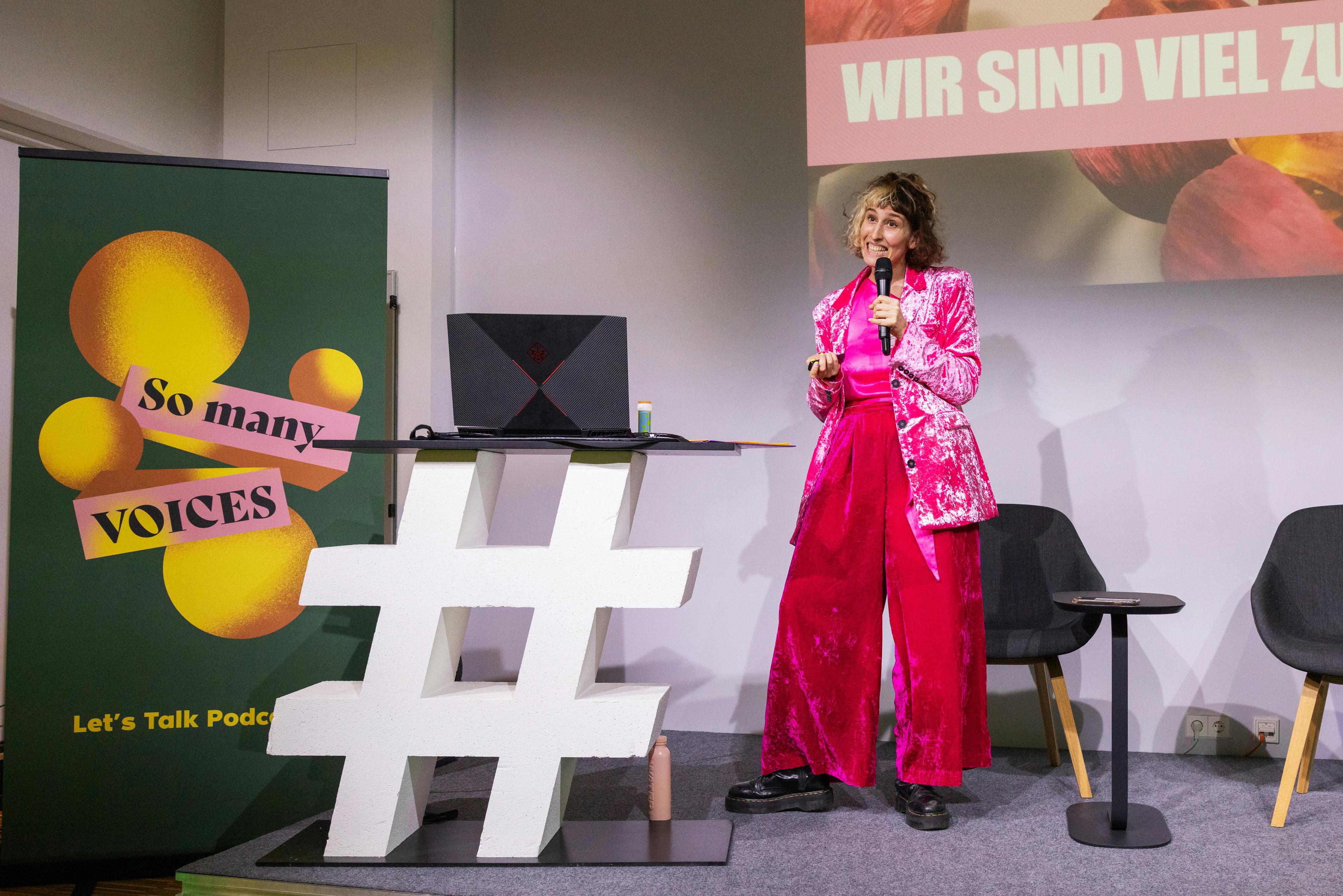 Jeanne Drach spricht auf der Bühne der So many Voices in ein Mikro. Jeanne trägt eine pinkes Samtoutfit. Links ist ein Aufsteller mit dem „So many Voices“ Logo. Im Hintergrund stehen zwei graue Sessel.