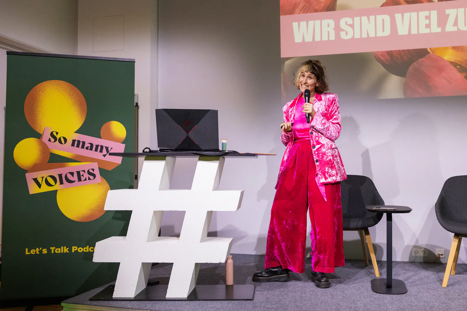 Jeanne Drach spricht auf der Bühne der So many Voices in ein Mikro. Jeanne trägt eine pinkes Samtoutfit. Links ist ein Aufsteller mit dem „So many Voices“ Logo. Im Hintergrund stehen zwei graue Sessel.