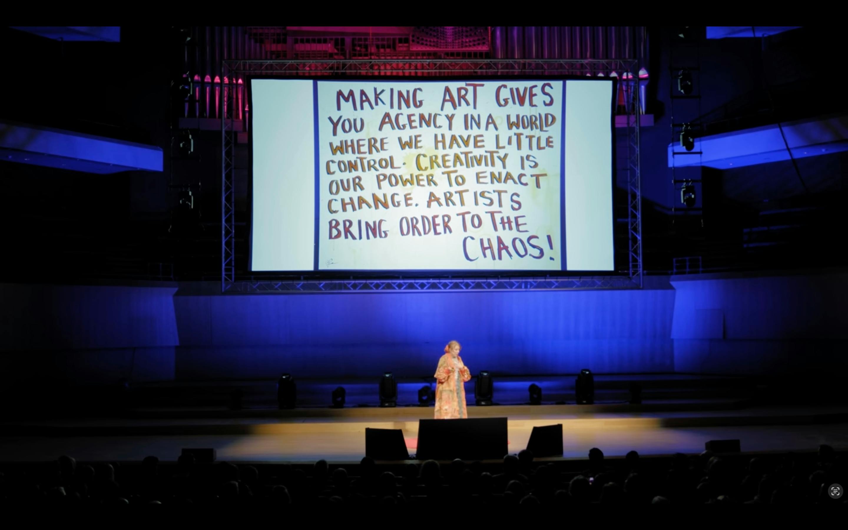 Screenshot aus dem TED-Talk von Amie McNee. Sie steht auf der Bühne. Hinter ihr ist eine große Leinwand, auf der folgendes steht: Making art gives you agency in a world where we have little control. Creativity is our power to enact change. Artists bring order to the chaos.