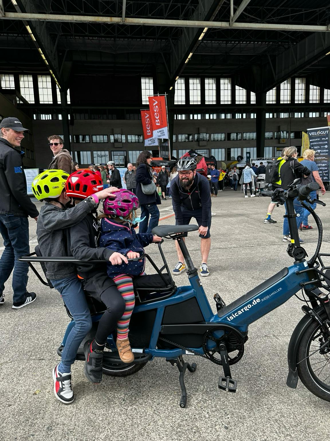 Drei Kinder hintereinander auf einem schmalen Lastenrad. Sie schauen ihren Papa an, daher sind ihre Gesichter nicht zu sehen.