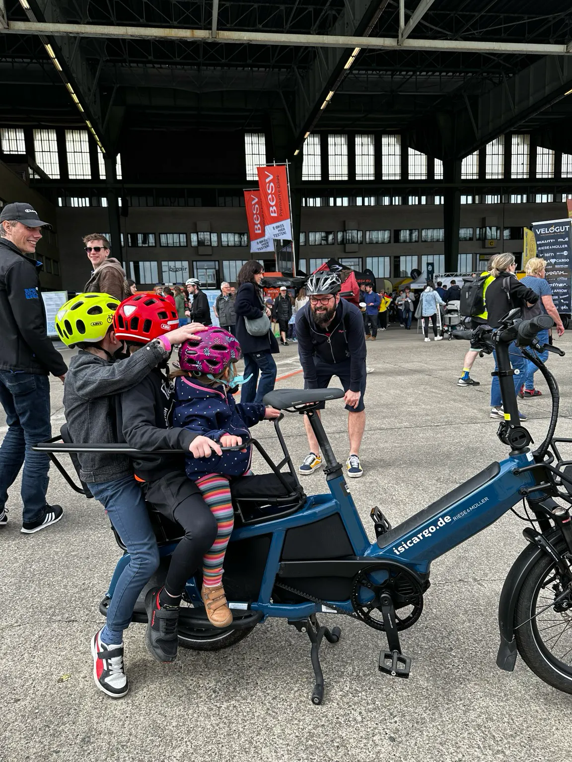Drei Kinder hintereinander auf einem schmalen Lastenrad. Sie schauen ihren Papa an, daher sind ihre Gesichter nicht zu sehen.