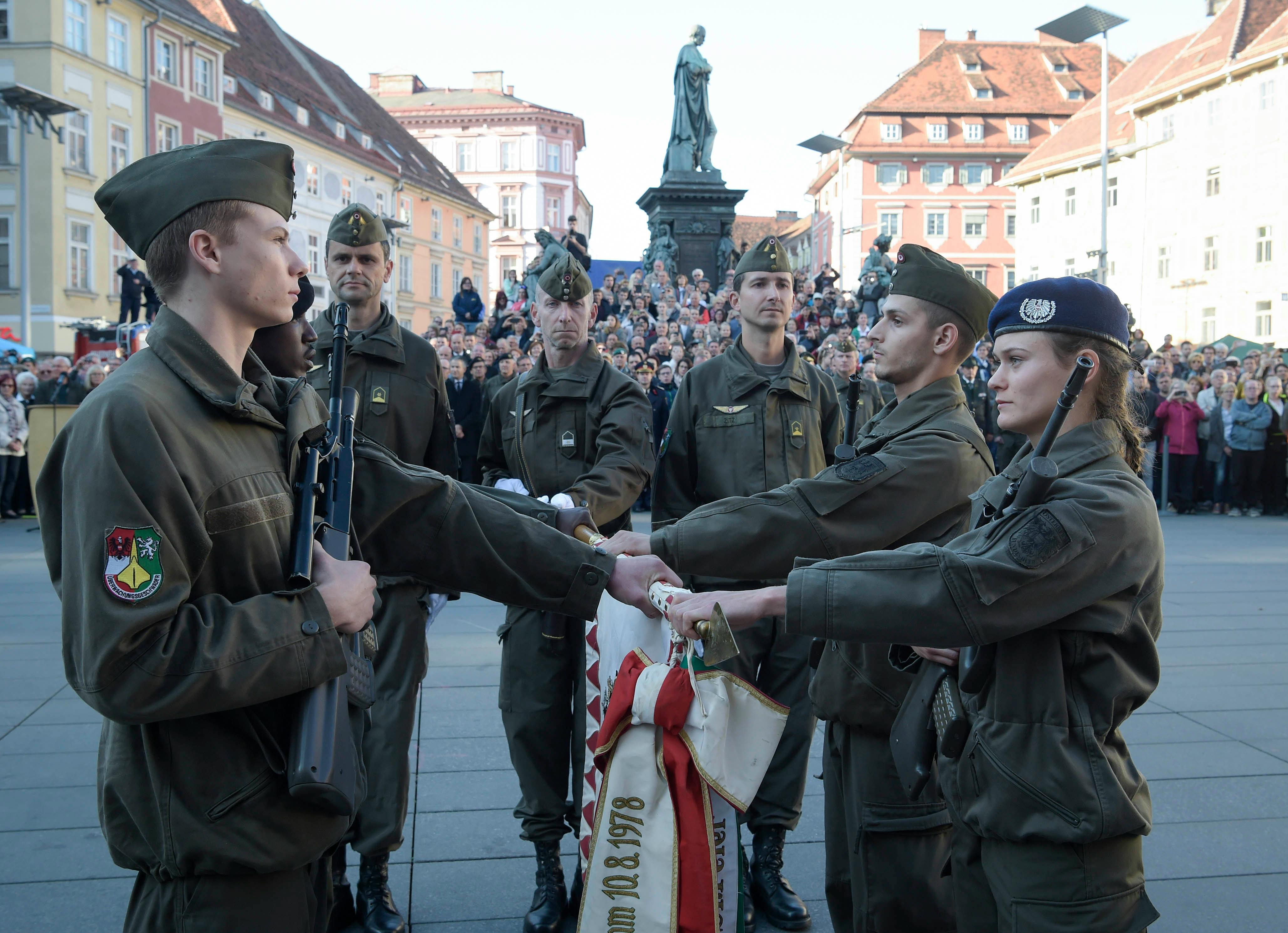 Das Bild zeigt eine Angelobung am Grazer Hauptplatz: Bundesheer-Soldatinnen und -Soldaten in olivgrüner Uniform legen gemeinsam die Hand auf die Truppenfahne. Im Hintergrund sind viele Zuschauer und historische Fassaden mit einer Statue zu sehen.