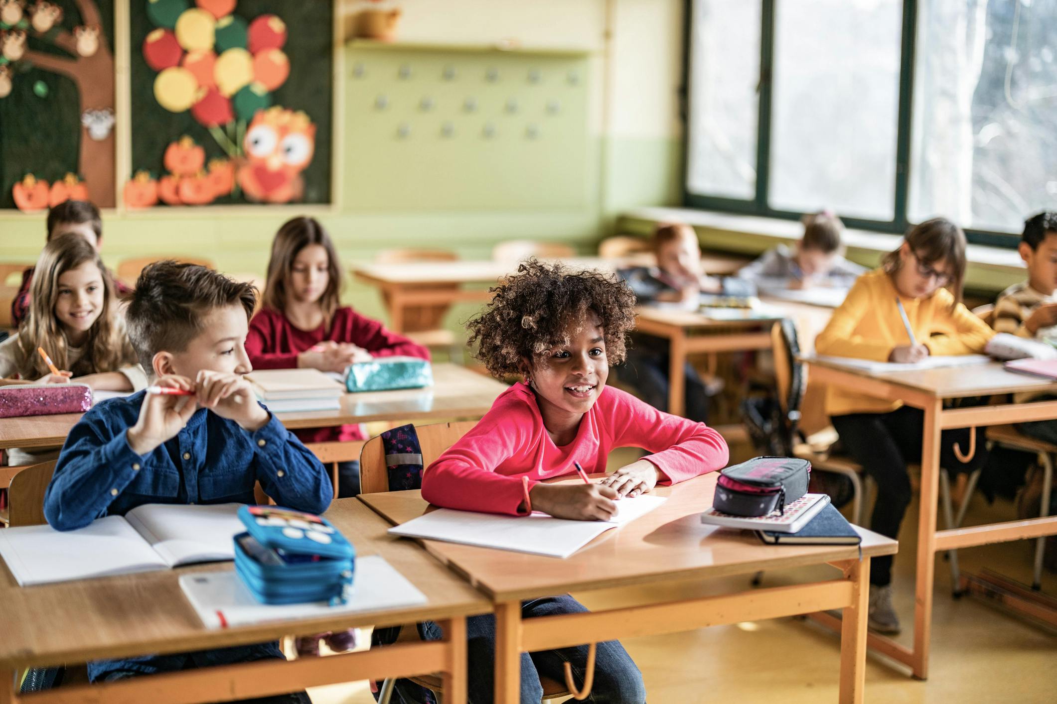 Dieses Foto zeigt Kinder in einem Klassenzimmer, die an ihren Schulbänken sitzen und arbeiten. Im Vordergrund lächelt ein Mädchen mit lockigen Haaren in die Kamera.