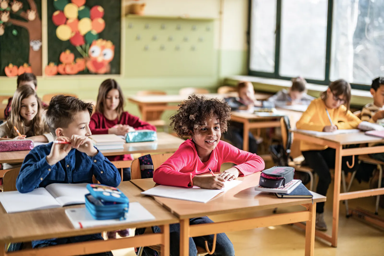 Dieses Foto zeigt Kinder in einem Klassenzimmer, die an ihren Schulbänken sitzen und arbeiten. Im Vordergrund lächelt ein Mädchen mit lockigen Haaren in die Kamera.