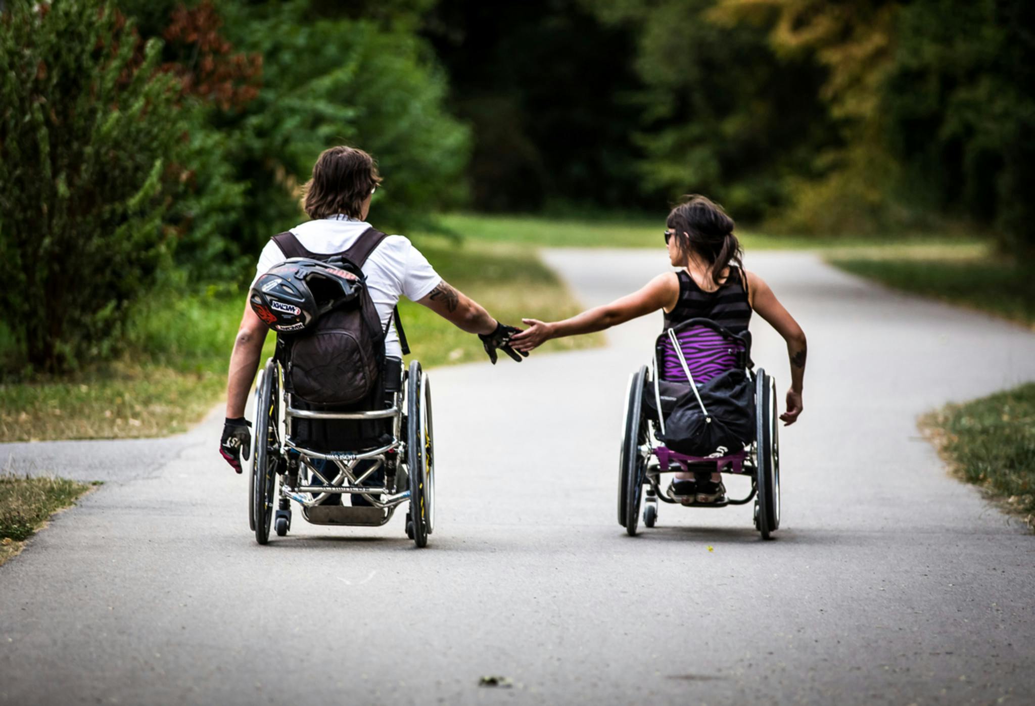 Zwei Menschen, die einen Rollstuhl nutzen, fahren auf einem geschlungenen Weg, rechts und links ist Parkfläche. Sie halten sich aus etwas Entfernung an den Händen.