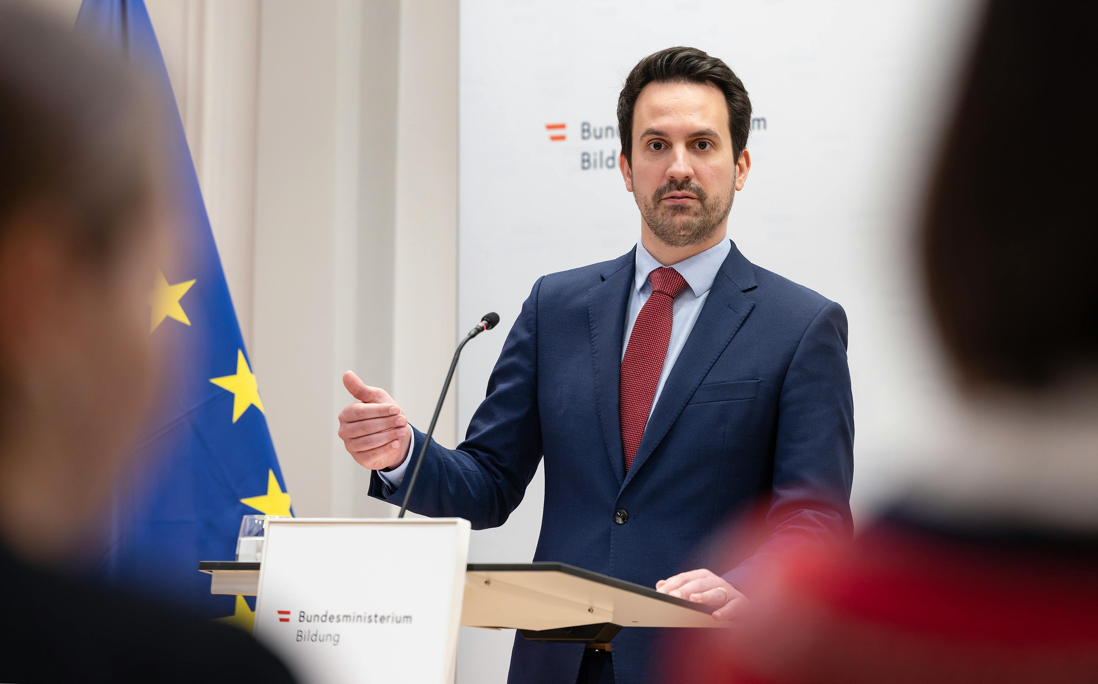Das Bild zeigt Bildungs-Minister Christoph Wiederkehr (NEOS) bei einer Pressekonferenz. Er steht an einem Rednerpult des Bundes-Ministeriums für Bildung und spricht mit einer Handgeste zu seinem Publikum. Im Hintergrund ist eine EU-Fahne zu sehen.