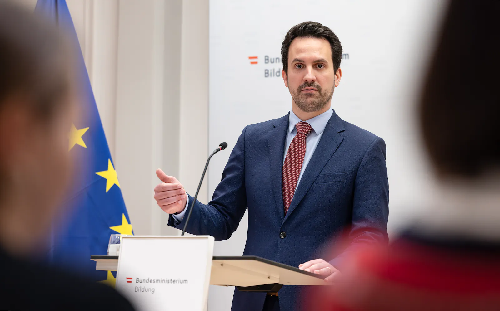 Das Bild zeigt Bildungs-Minister Christoph Wiederkehr (NEOS) bei einer Pressekonferenz. Er steht an einem Rednerpult des Bundes-Ministeriums für Bildung und spricht mit einer Handgeste zu seinem Publikum. Im Hintergrund ist eine EU-Fahne zu sehen.