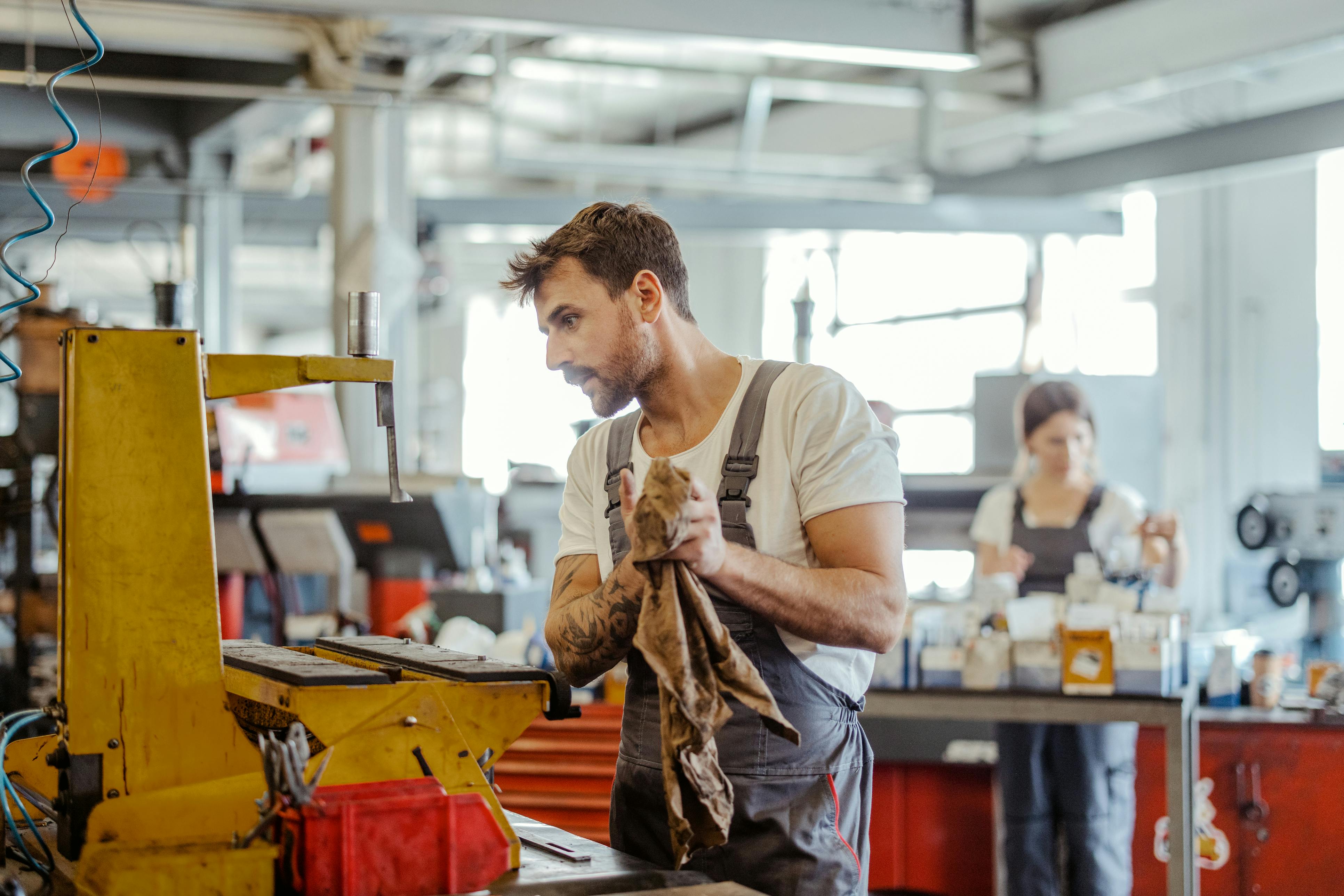 Das Bild zeigt einen Mann in Arbeits-Kleidung in einer Industrie-Halle. Im Hintergrund arbeitet eine Frau an einer Maschine.