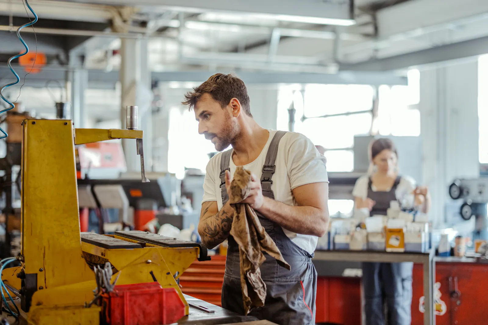 Das Bild zeigt einen Mann in Arbeits-Kleidung in einer Industrie-Halle. Im Hintergrund arbeitet eine Frau an einer Maschine.