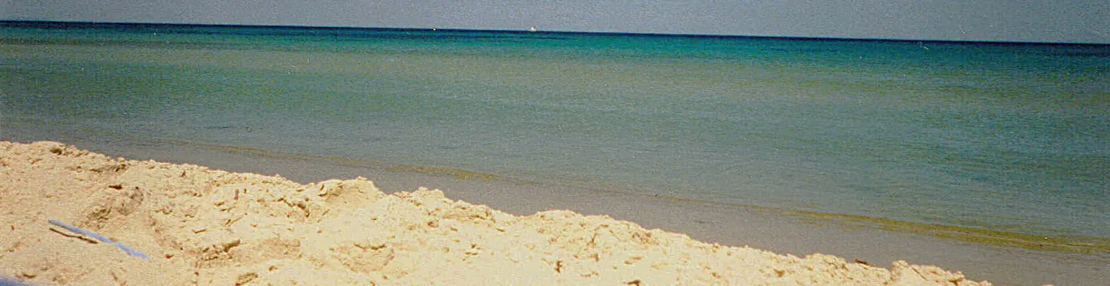 Kontrastreiches Bild: Im Vordergrund beiger Sandstrand, diagonal im Hintergrund das blaue Meer mit dunkelblauem Horizont.