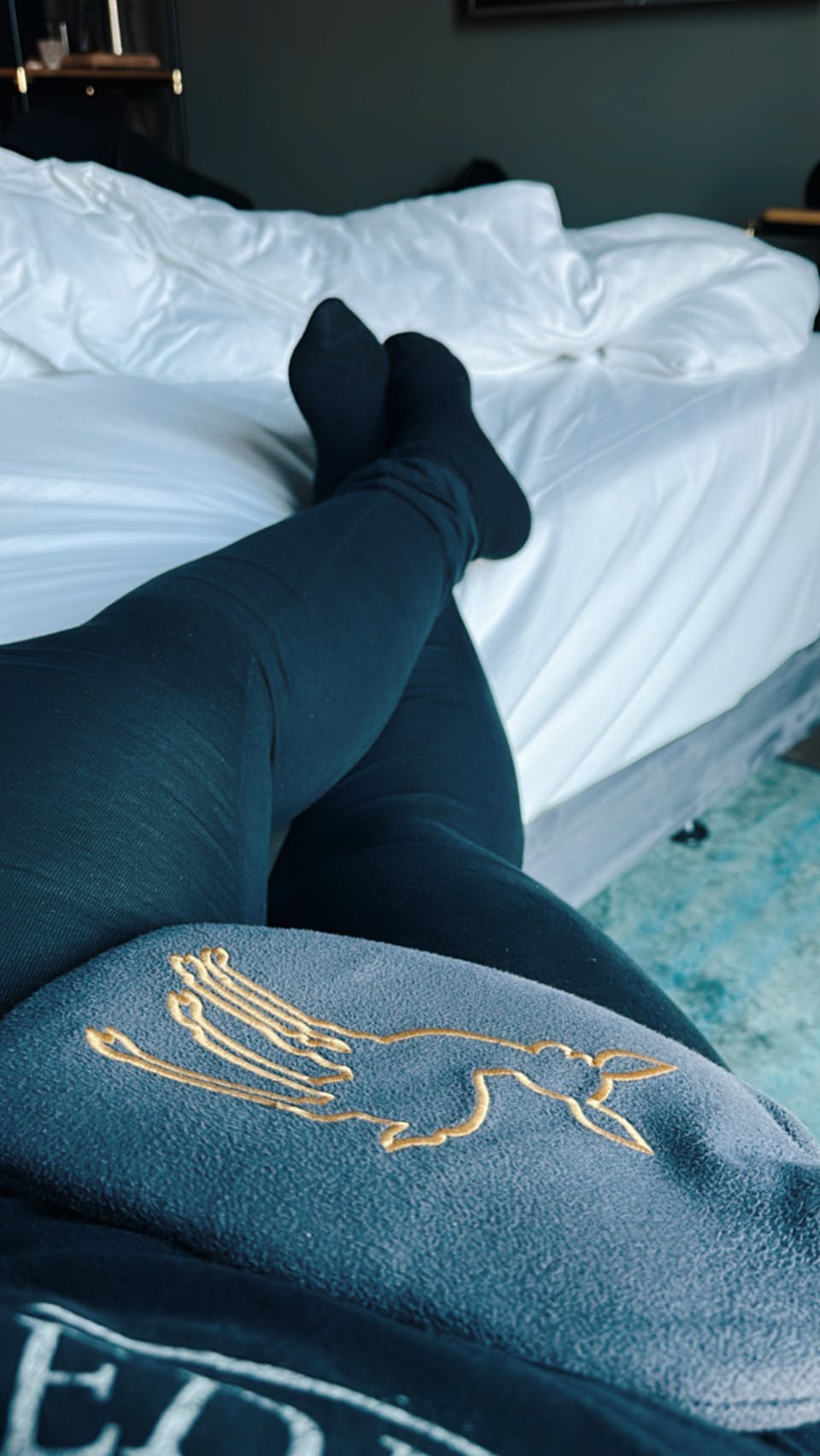 Auf einem Bett hochgelegte Beine in Leggings und Strümpfen, auf dem Bauch liegt eine Wärmflasche.