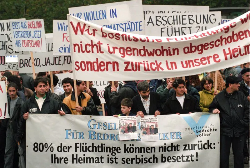 foto einer demo gegen den sogenannten "asylkompromiss" 1993. auf dem foto sind bosnische flüchtlinge mit transparenten. Auf dem größten, ganz vorne, steht "80% der flüchtlinge können nicht zurück: ihre heimat ist serbisch besetzt!" direkt dahinter steht auf einem anderen transparent "wir bosnischen Flüchtlinge wollen nicht irgendwohin abgeschoben werden, sondern zurück in unsere heimat". Und dahinter hält jemand ein plakat mit den worten "Erst aus bijeljina vertrieben, jetzt aus Deutschland!"