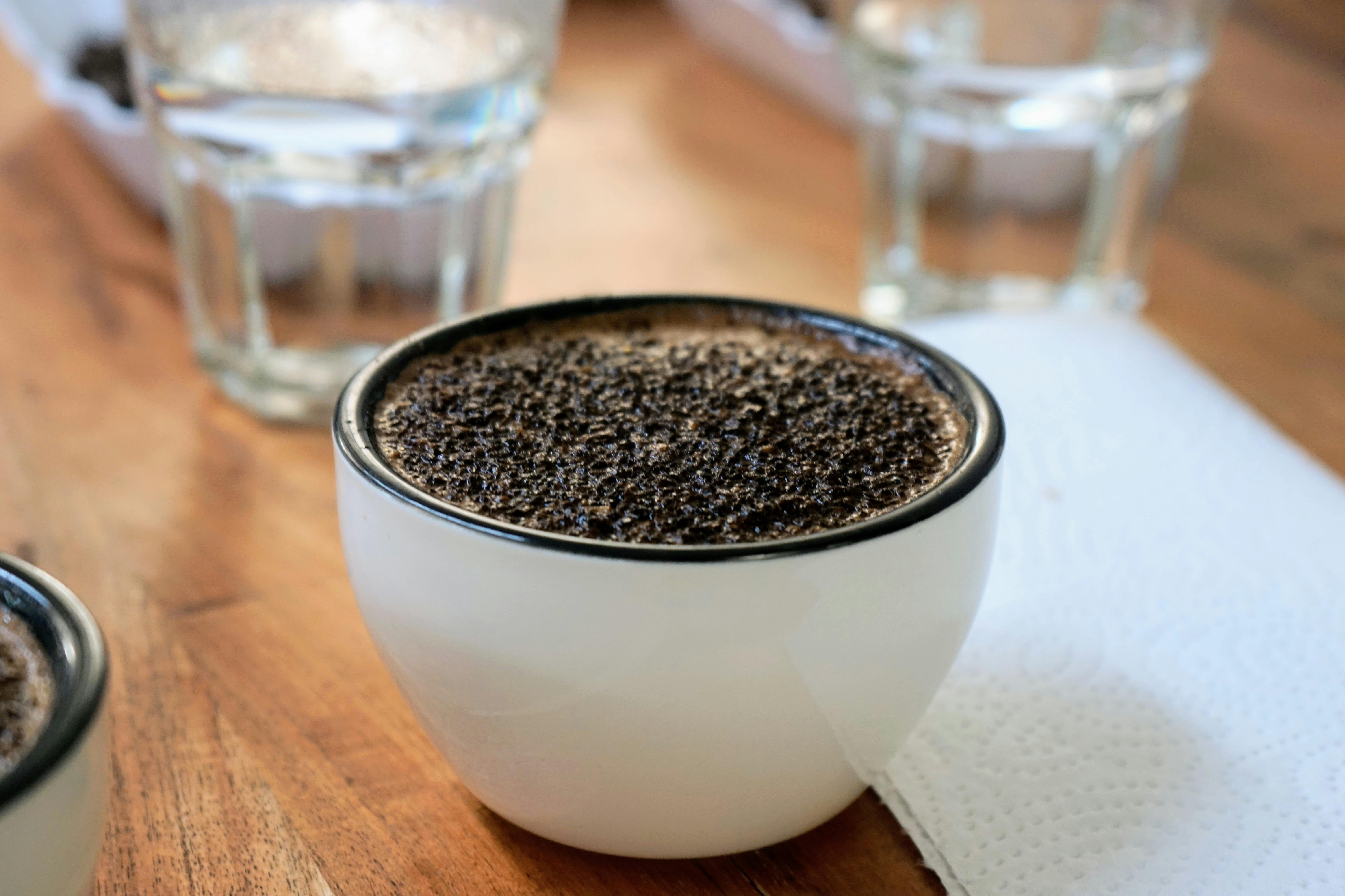 Makroaufnahme einer weißen Kaffeetasse, auf der eine dunkle, aromatische Kaffeekruste aus grobem Kaffeemehl schwimmt. Text: Die Schritt-für-Schritt-Anleitung für das perfekte Cupping zu Hause.