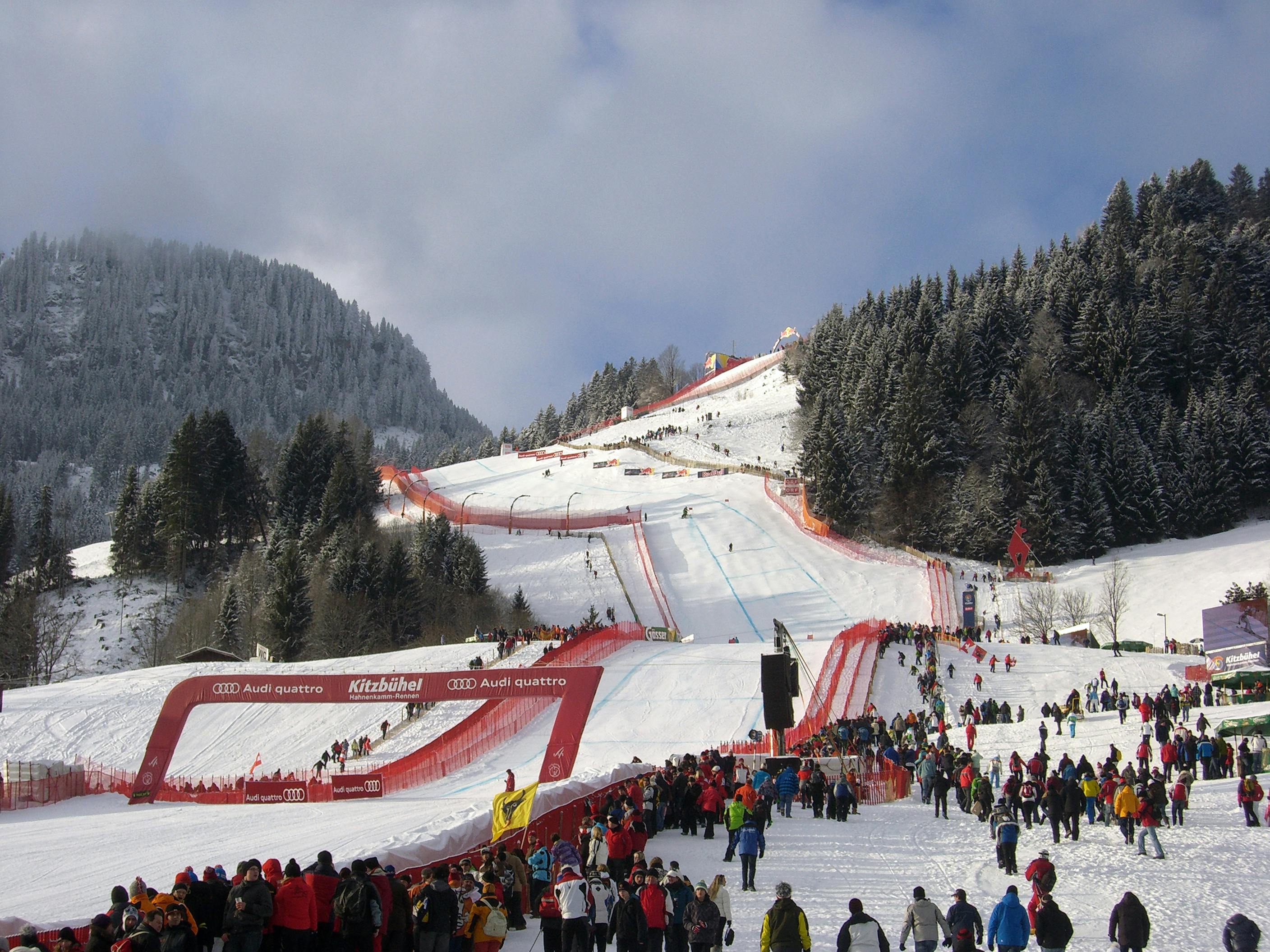Das Bild zeigt einen Blick auf den Zielbereich der Streif in Kitzbühel. Die Skipiste ist mit roten Sicherheitsnetzen gesäumt. Hunderte Zuschauer in bunten Winterjacken stehen am Rand der Piste. Im Hintergrund sind verschneite Berge und Wälder zu sehen. Eine große Tribüne ist mit Menschen gefüllt.