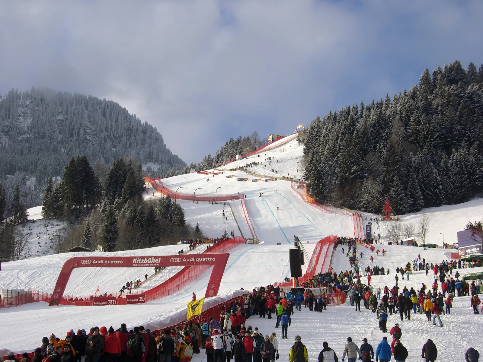 Das Bild zeigt einen Blick auf den Zielbereich der Streif in Kitzbühel. Die Skipiste ist mit roten Sicherheitsnetzen gesäumt. Hunderte Zuschauer in bunten Winterjacken stehen am Rand der Piste. Im Hintergrund sind verschneite Berge und Wälder zu sehen. Eine große Tribüne ist mit Menschen gefüllt.