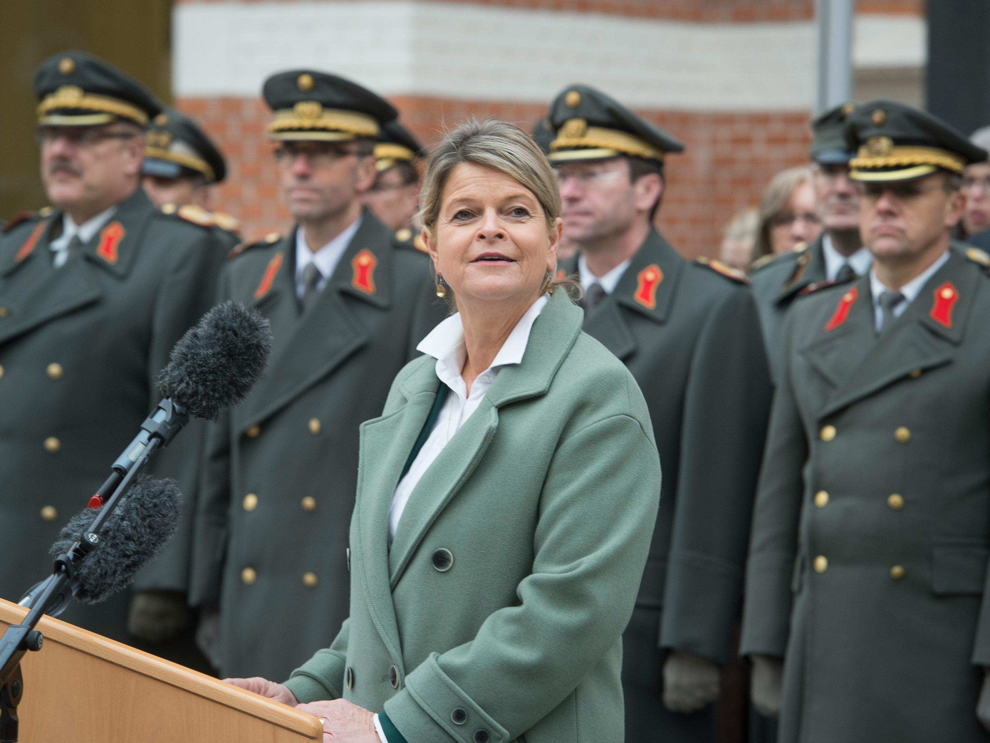 Verteidigungsministerin Klaudia Tanner steht in einem grünen Blazer an einem Rednerpult mit Mikrofonen. Im Hintergrund stehen Offiziere des österreichischen Bundesheers in Uniformen mit goldenen Schulterklappen.