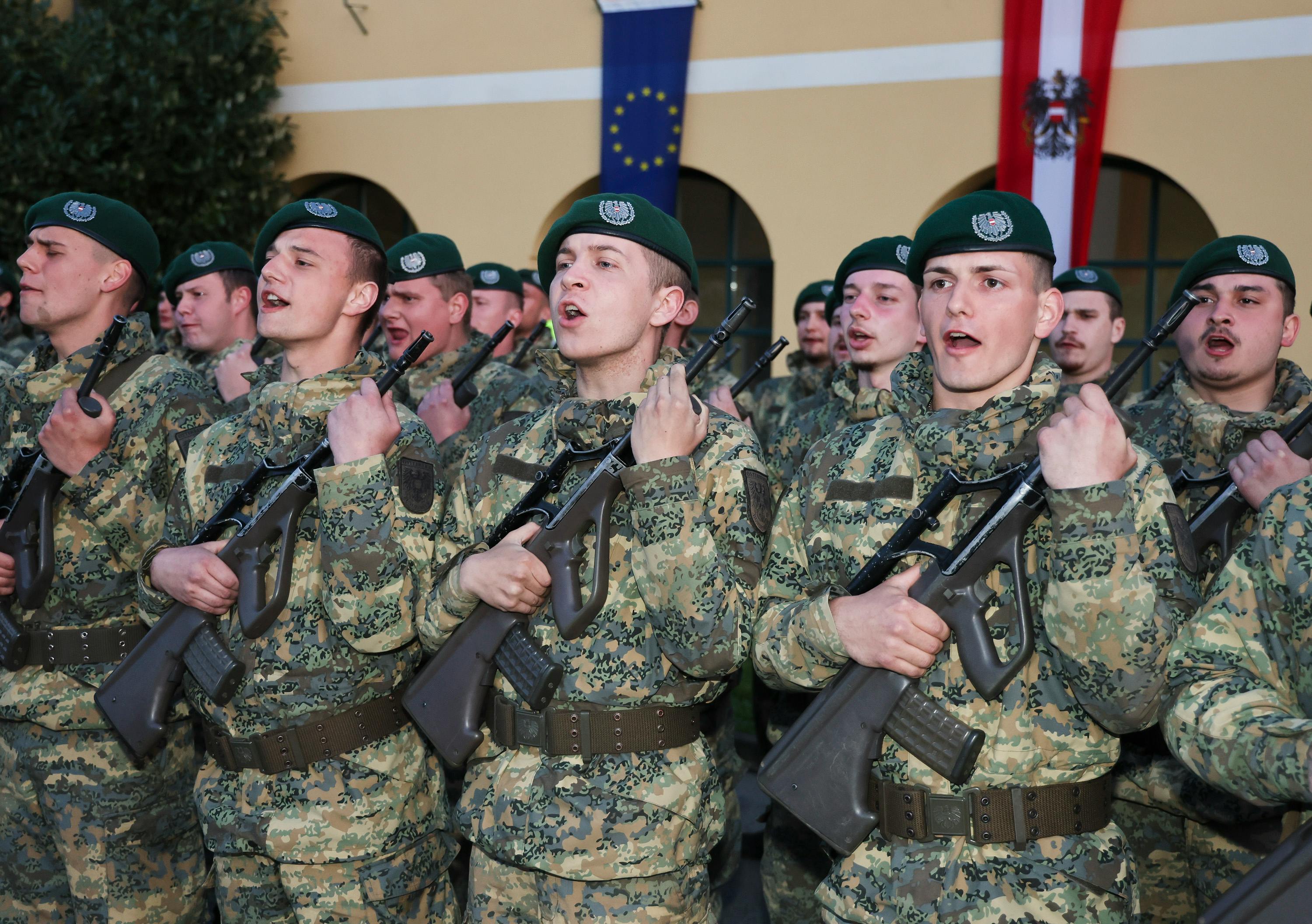 Junge Soldaten des österreichischen Bundesheers in Tarnuniformen und grünen Baretts stehen in Formation und legen den Schwur ab. Sie halten Gewehre und heben ihre rechte Hand. Im Hintergrund sind die EU-Flagge und die österreichische Flagge zu sehen.