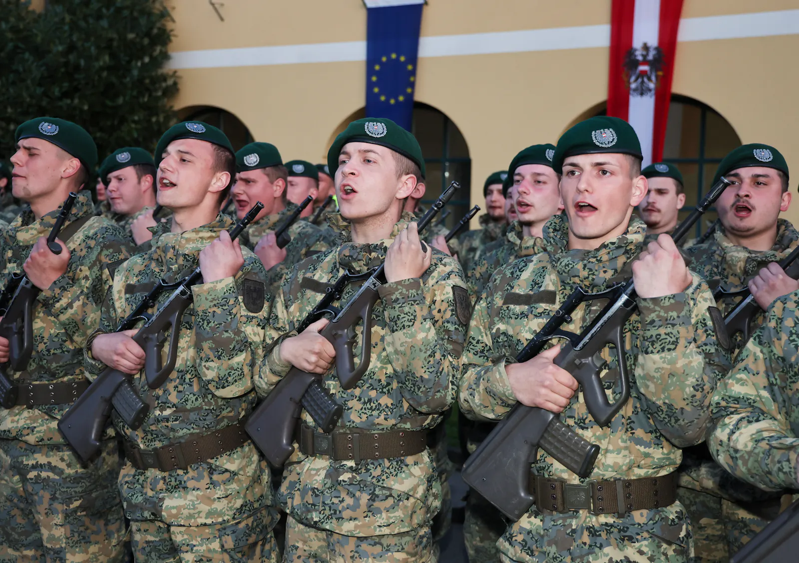 Junge Soldaten des österreichischen Bundesheers in Tarnuniformen und grünen Baretts stehen in Formation und legen den Schwur ab. Sie halten Gewehre und heben ihre rechte Hand. Im Hintergrund sind die EU-Flagge und die österreichische Flagge zu sehen.
