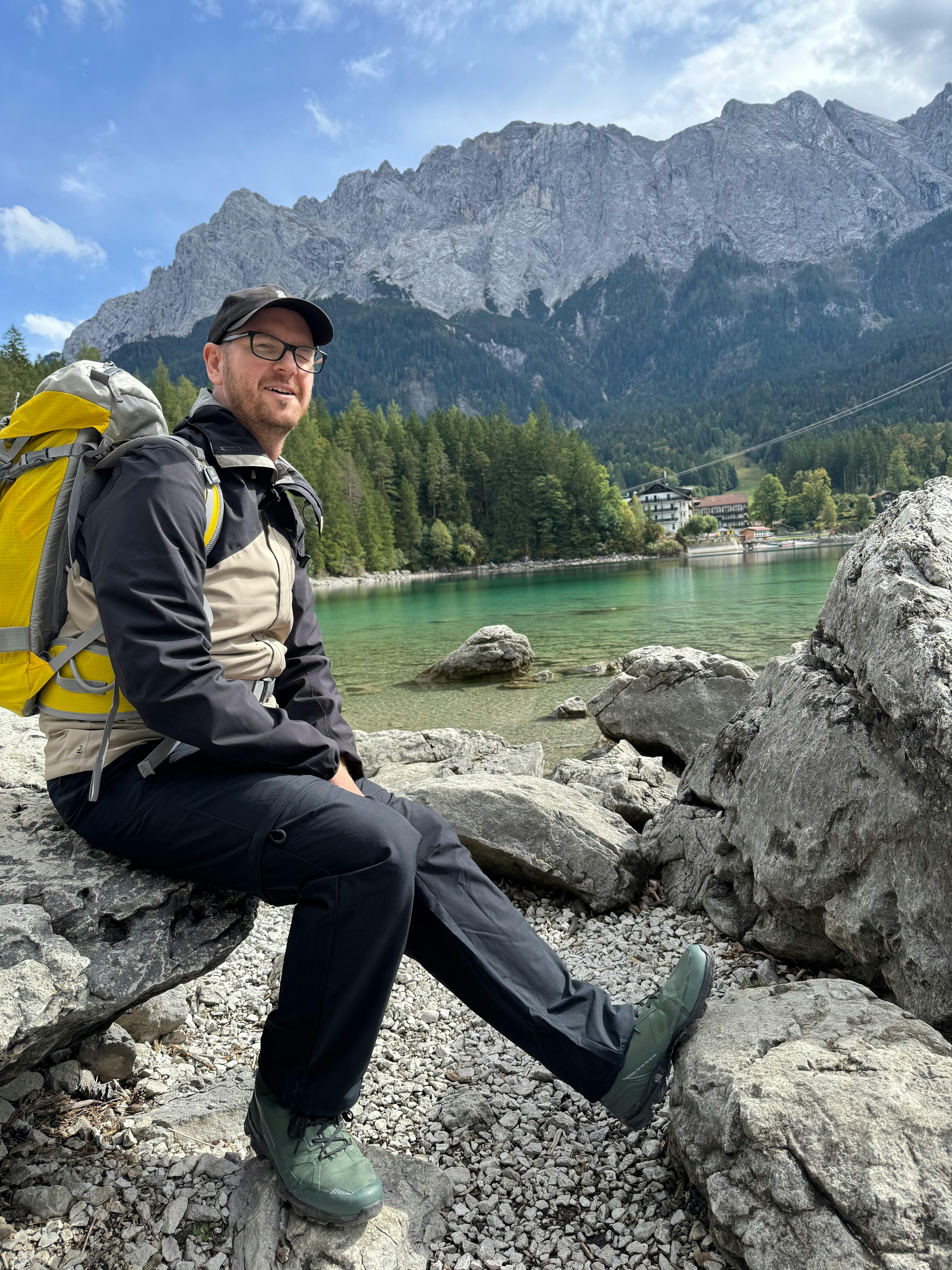 Christian sitzt mit Wanderrucksack vor der Zugspitze an einem See.