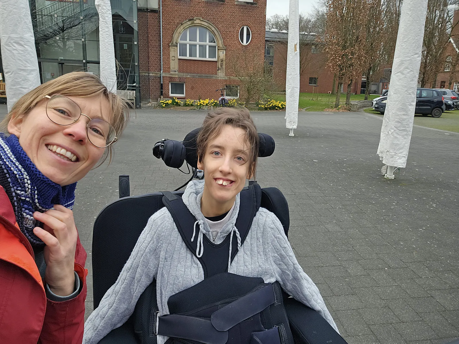 Ein Selfie von 2 Personen, die in die Kamera lachen. Eine Frau mit roter Jacke hält die Kamera. Die zweite jüngere Frau hat eine Behinderung und sitzt in einem Elektrorollstuhl.