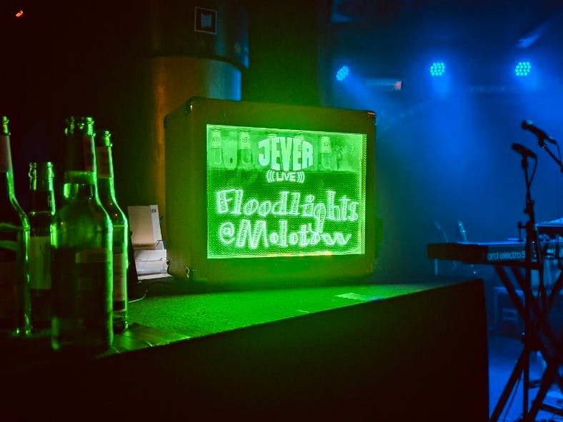 Leere Flaschen, ein grün leuchtendes Schild mit der Konzerntankündigung "Floodlights @ Molotow" und Teile der Bühne mit Mikrofonen