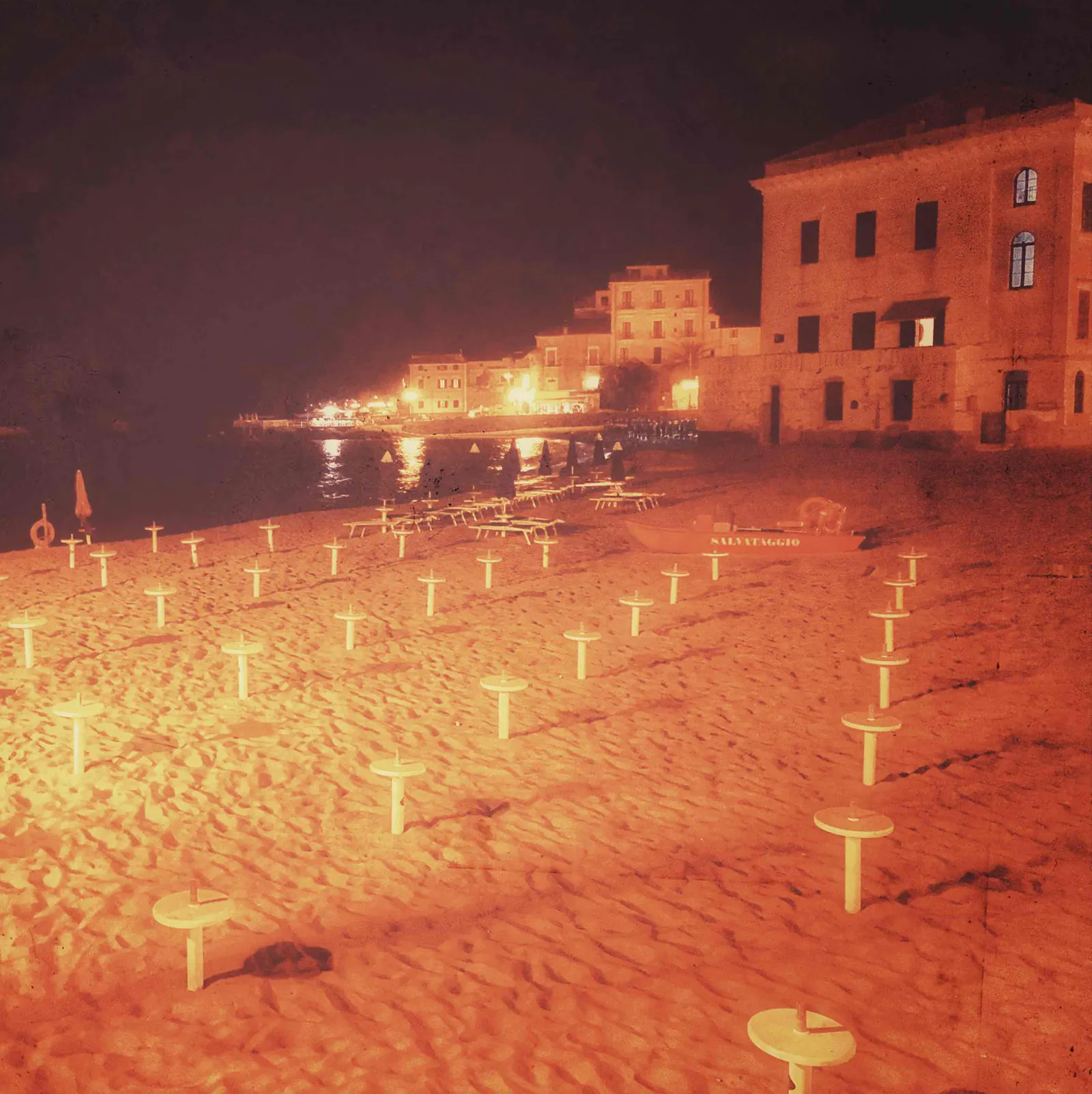 Blick auf einen menschenleeren, gepflegten Strand, mit Stümpfen abgebauter Sonenschirme. Im Hintergrund Häuser mit teils erleuchteten Fenstern und das ruhige Meer.