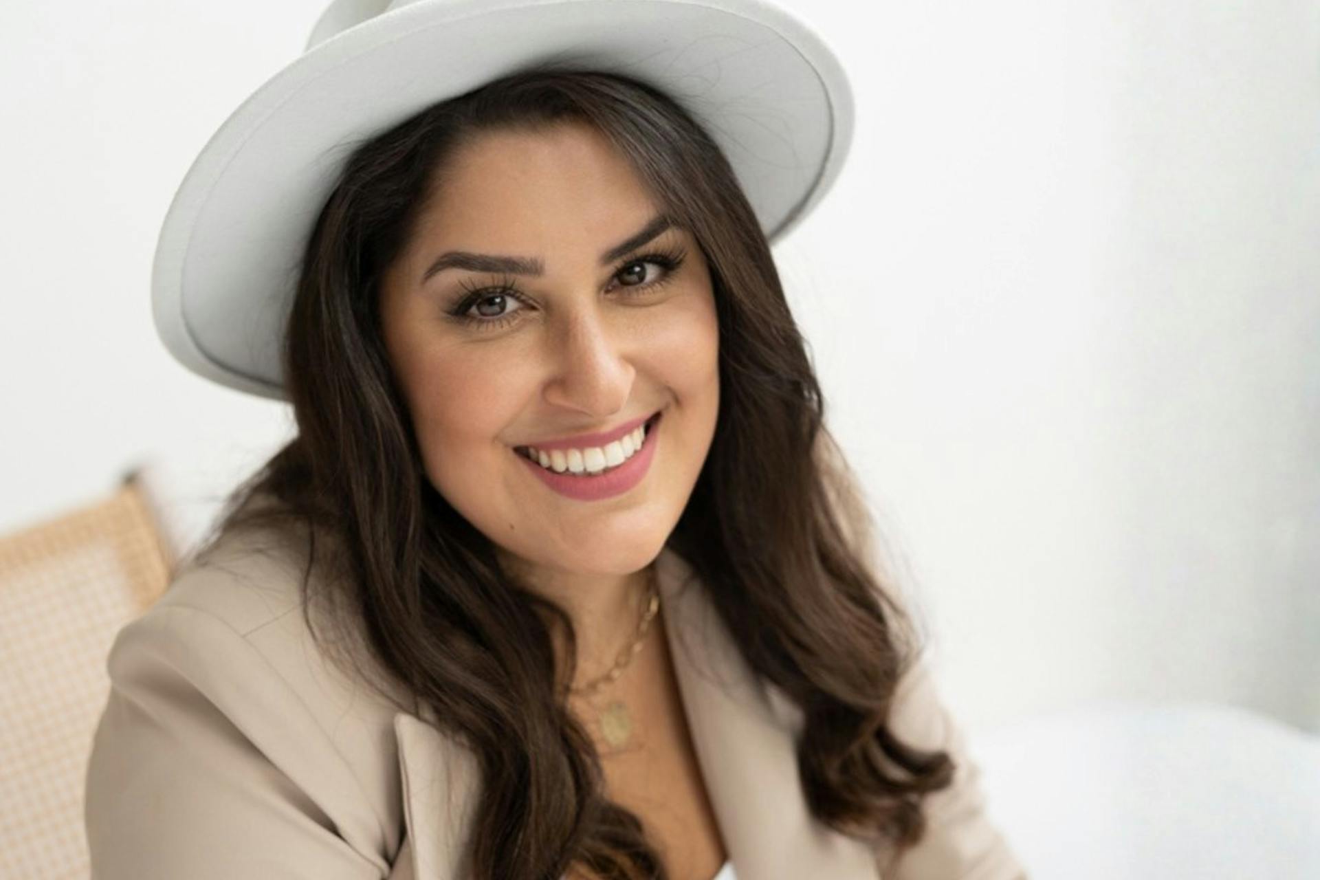 Duygu Bayramoglu trägt einen beigefarbenen Blazer und einen weißen Hut und sitzt in einem weißen Raum. Sie lächelt.
