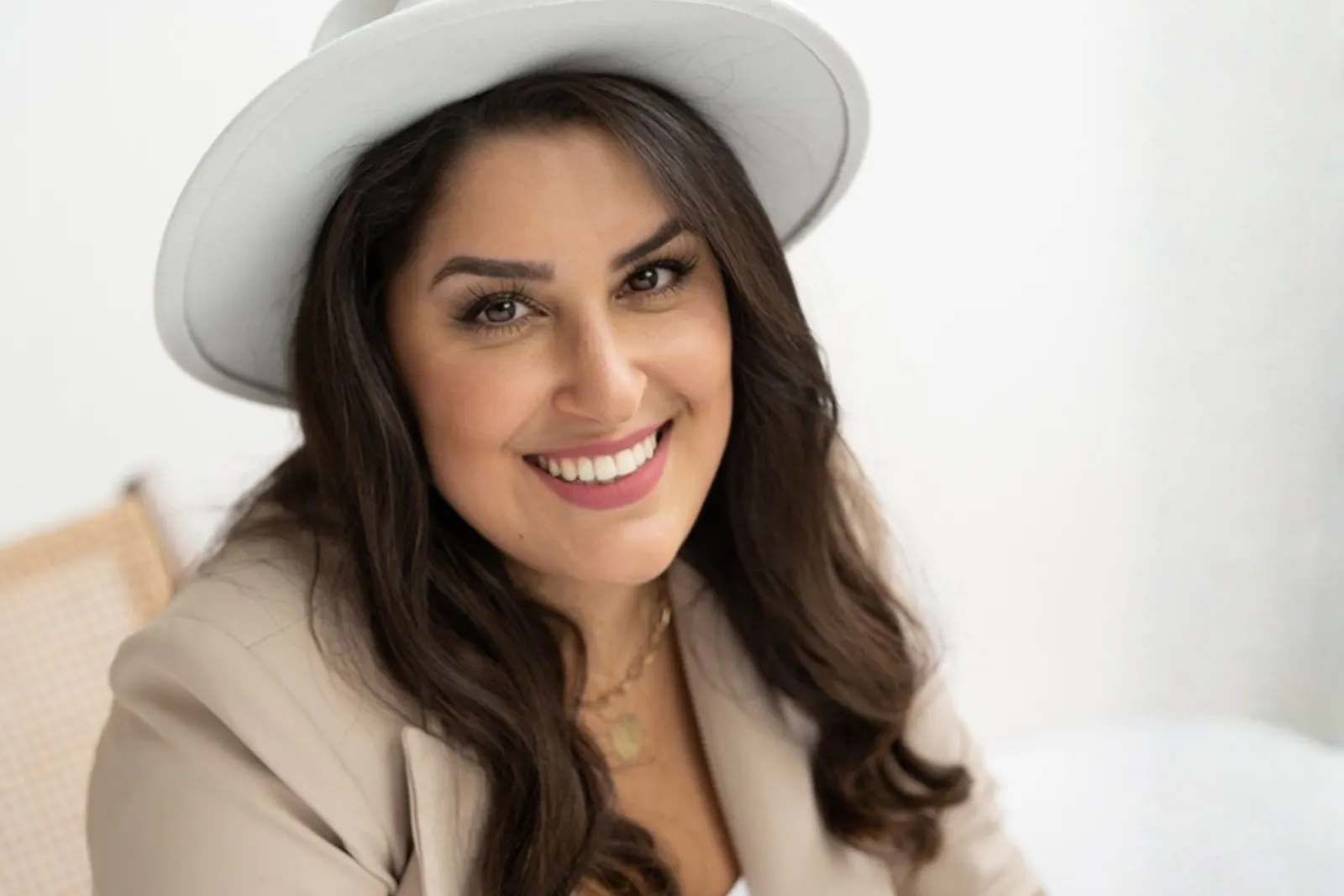 Duygu Bayramoglu trägt einen beigefarbenen Blazer und einen weißen Hut und sitzt in einem weißen Raum. Sie lächelt.