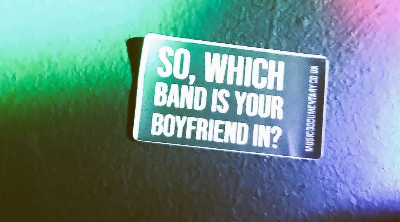 Ein Aufkleber an einer Wand, von bunten Lichtern erleuchtet. Auf dem Aufkleber steht: "So, which band is your boyfriend in?"
