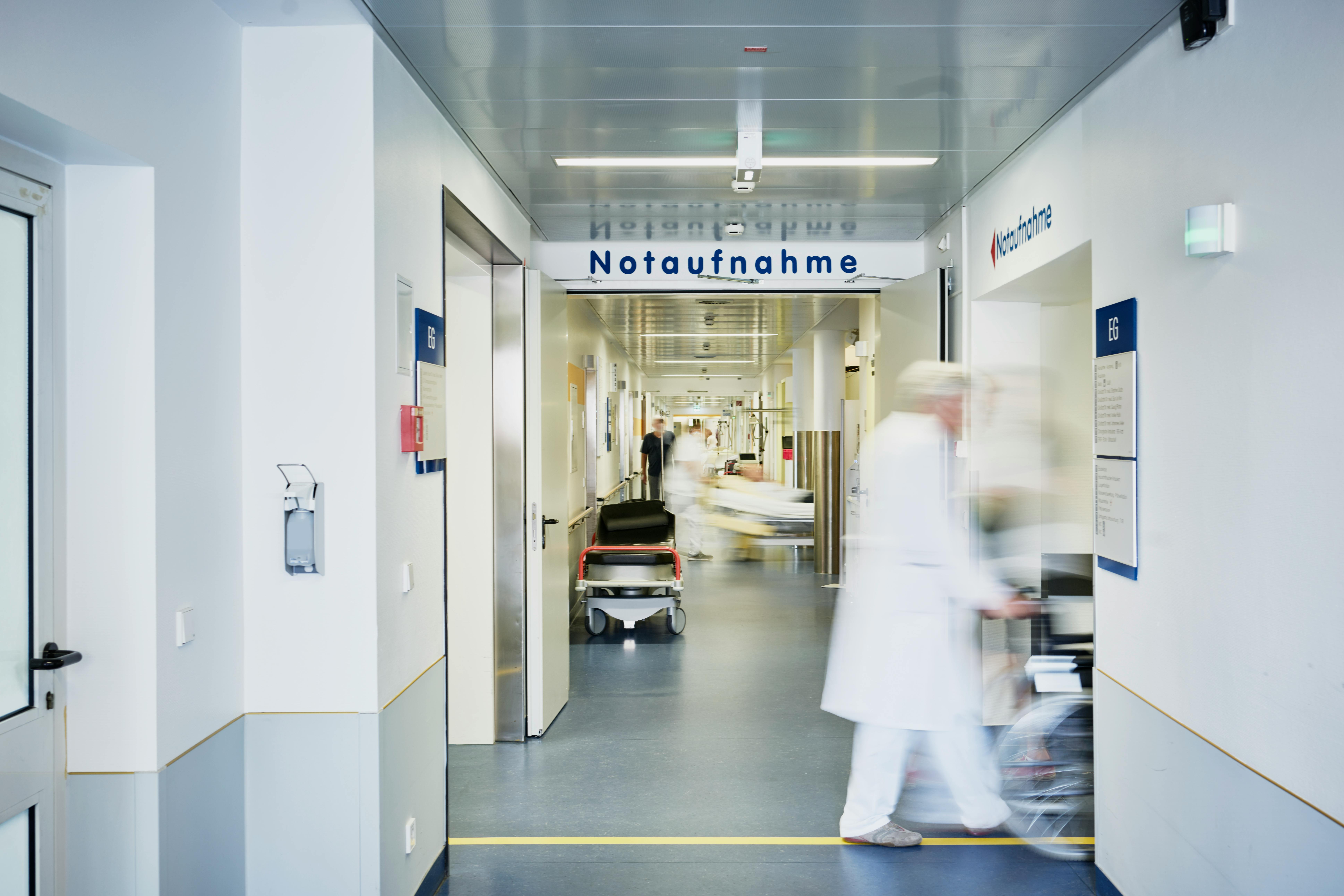 Das Bild zeigt einen Flur einer Notaufnahme mit Schild „Notaufnahme“, einen unscharfen Mitarbeitenden und leerer Trage.